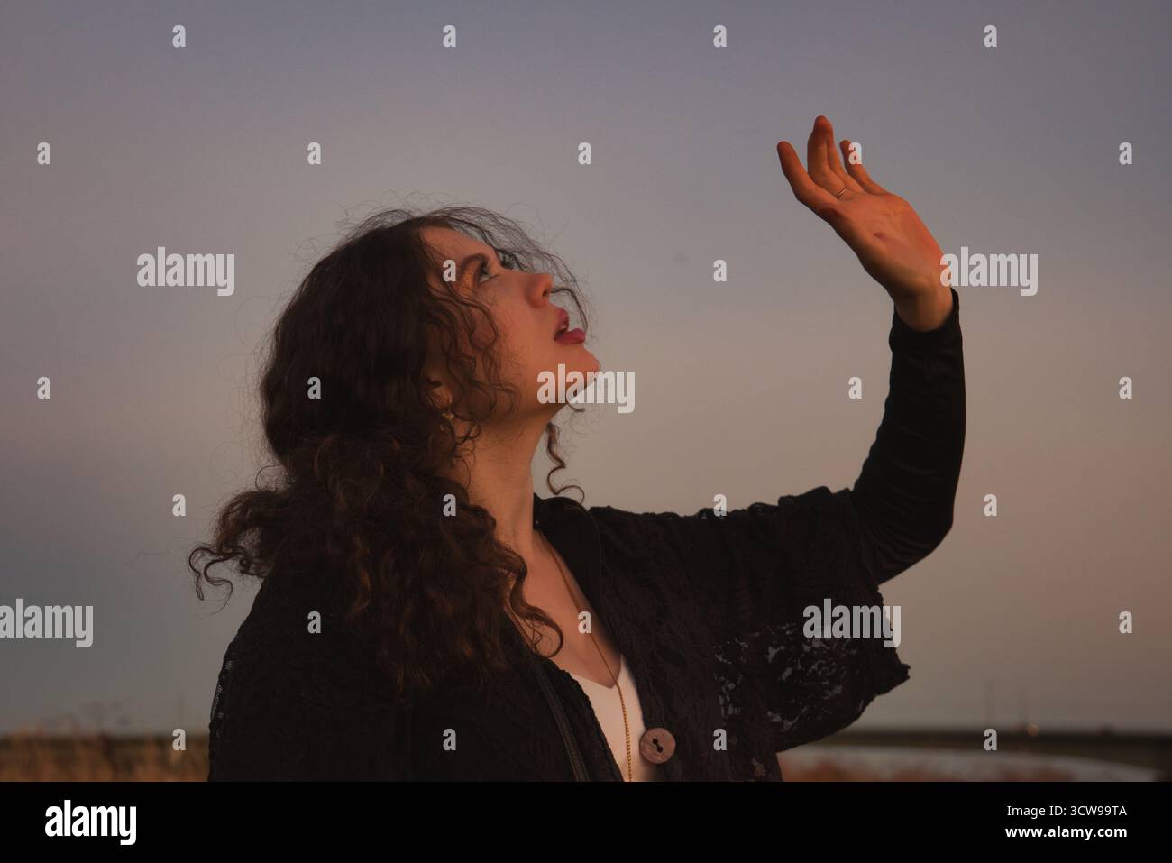 Donna ispiratrice che punta la mano verso l'alto verso Sunset Sky con capelli ricci e posa espressiva per motivi libertà e temi di aspirazione in outdo Foto Stock