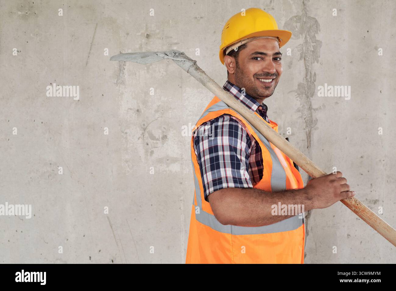 Ritratto di un giovane uomo mediorientale che sorride tenendo la pala sulla spalla, indossando casco da costruzione e giubbotto di sicurezza, in piedi contro un muro di cemento infinito Foto Stock