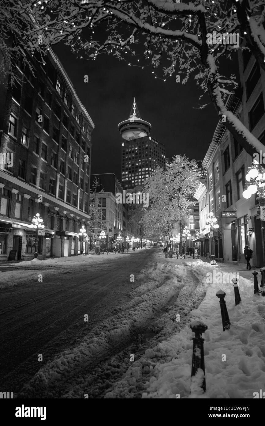 Una strada tranquilla è coperta di neve di notte, con luci scintillanti sugli alberi. Gli alti edifici circondano la zona e la gente passeggia lungo il sidewa innevato Foto Stock