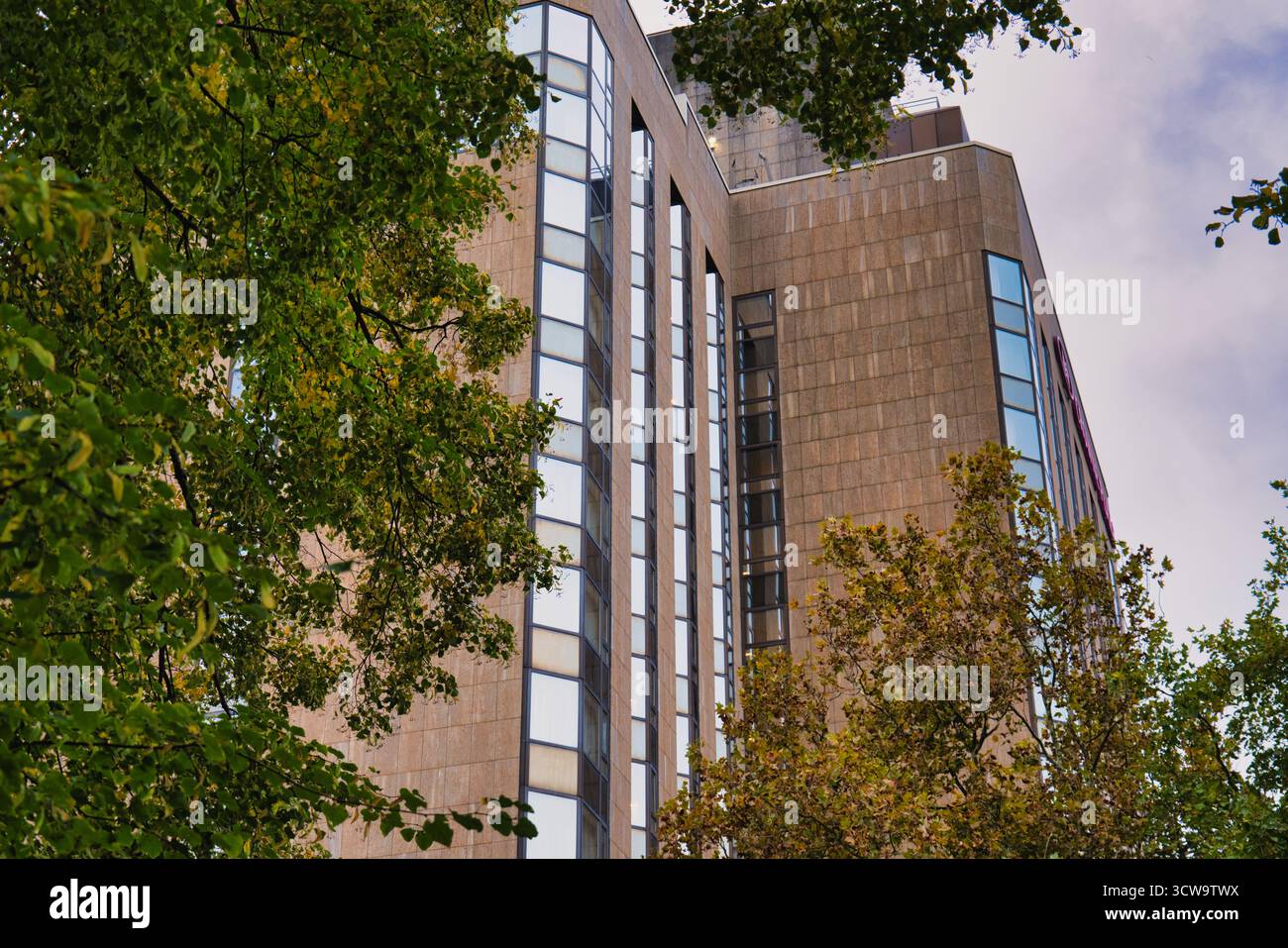 Alto e moderno edificio dietro alberi verdi, architettura urbana con finestre in vetro nel centro città, contrasto naturale e architettonico in autunno Foto Stock