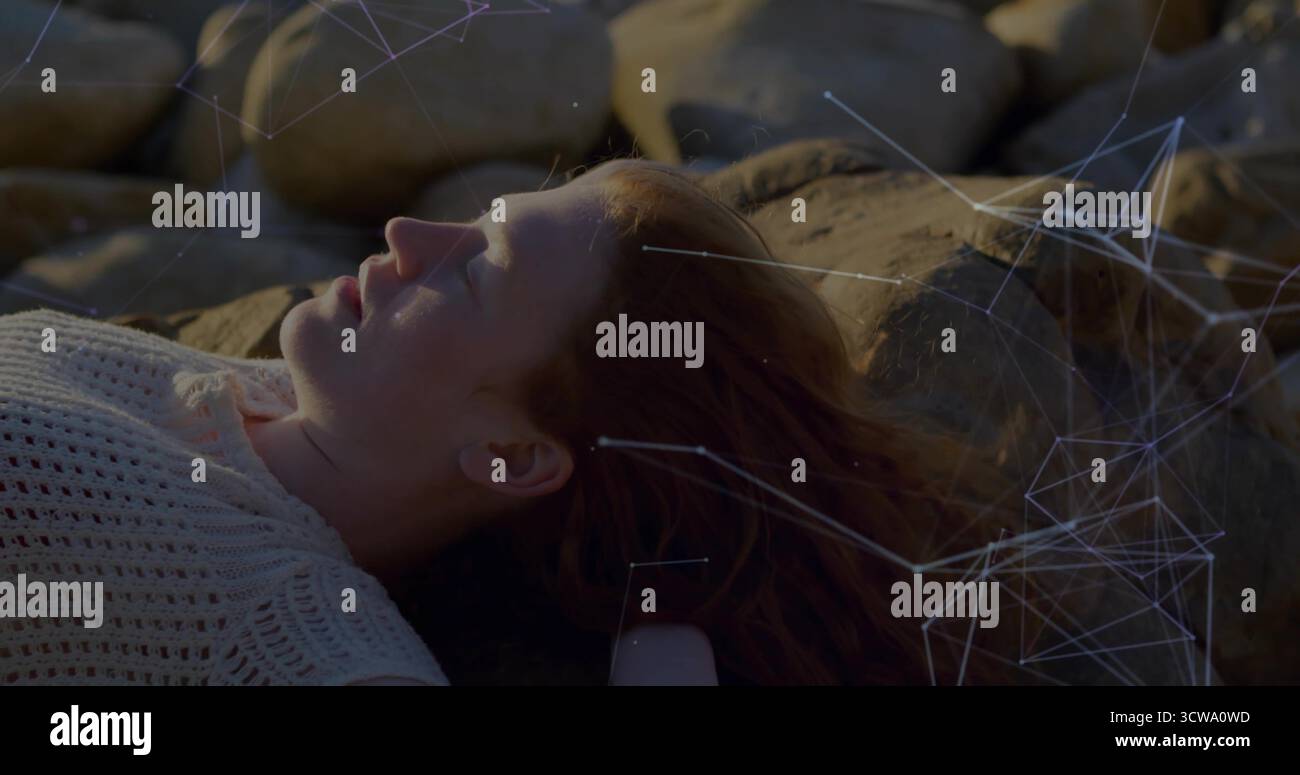 Donna sdraiata che indossa la parte superiore bianca sulla costa rocciosa, con rete luminosa e contenitore sotto i capelli Foto Stock