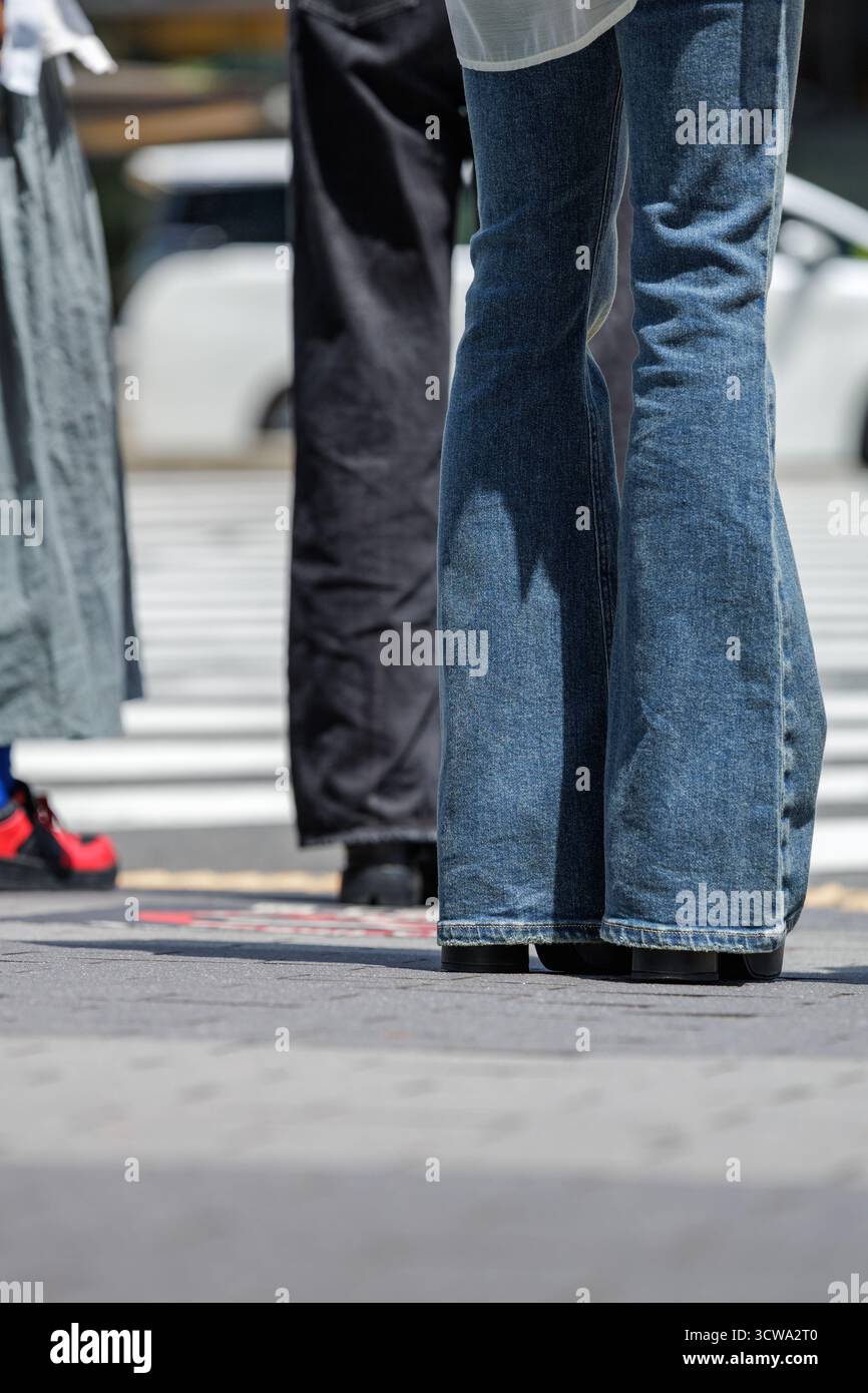 Jeans con fondo a campana e scarpe con plateau che ricordano la moda degli anni '1970 adornano una giovane donna giapponese che aspetta un incrocio zebra in un ambiente urbano frenetico. Foto Stock