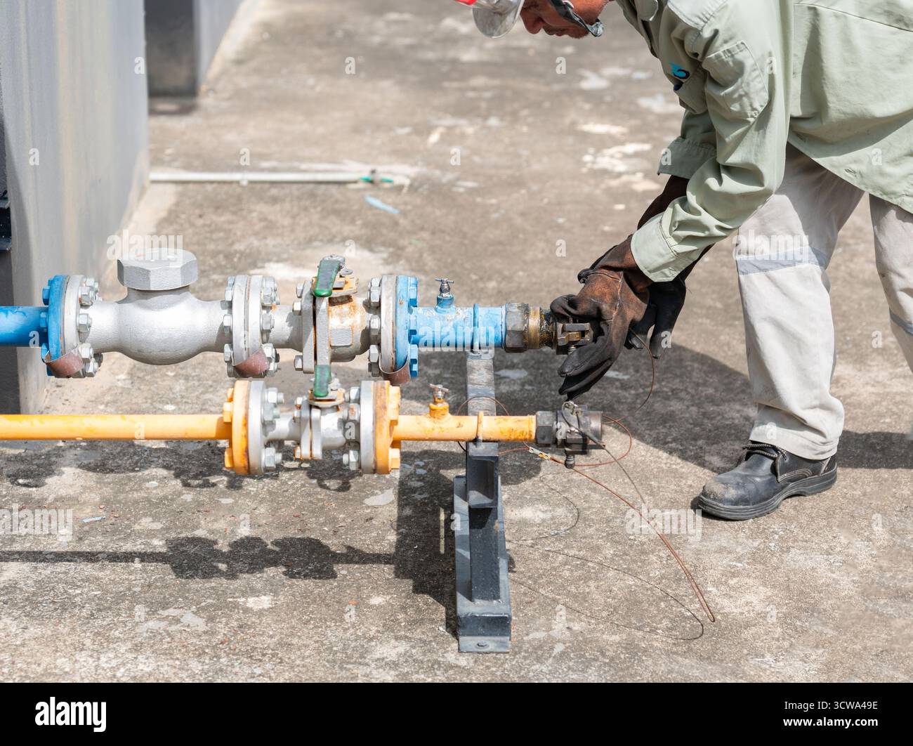 Il lavoratore è vestito con indumenti protettivi, tra cui guanti, stivali e un casco, mentre regola una valvola su un sistema di tubazioni del gas con codice colore. Foto Stock