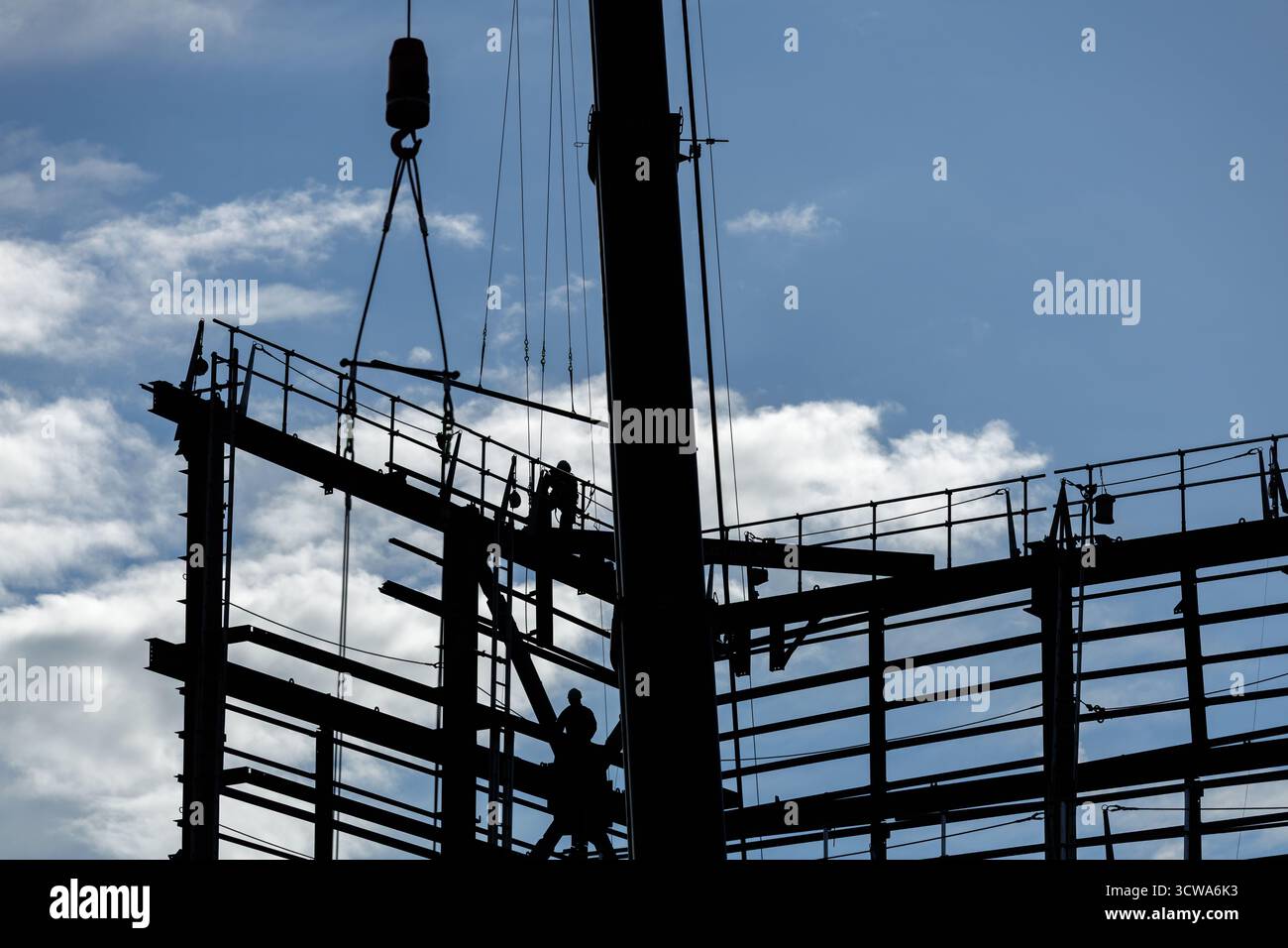 Ambiente industriale con lavoratori in attesa di sollevare il carico con gru mentre si trova sulla struttura dell'edificio, sagomata contro un cielo blu brillante con nuvole sparse. Foto Stock