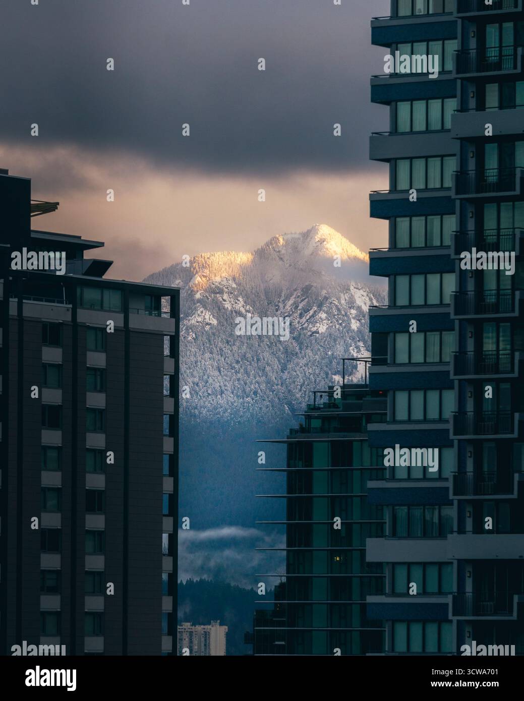 Le vette innevate si innalzano drammaticamente tra gli edifici moderni di una città, mostrando un bellissimo contrasto tra natura e architettura durante la serata Foto Stock