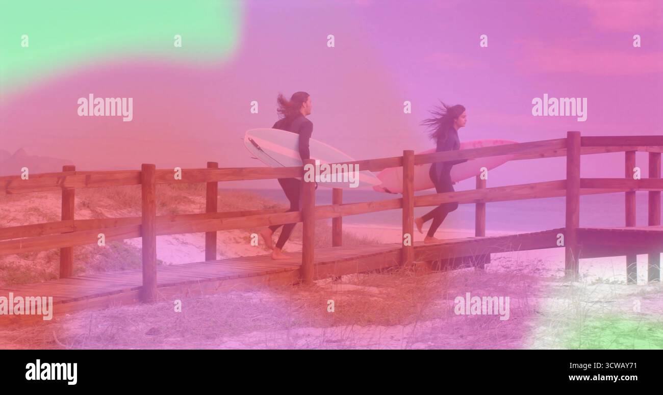 Coppia che cammina indossando muta e trasportando tavole da surf sul lungomare con erbe dune verso l'oceano Foto Stock