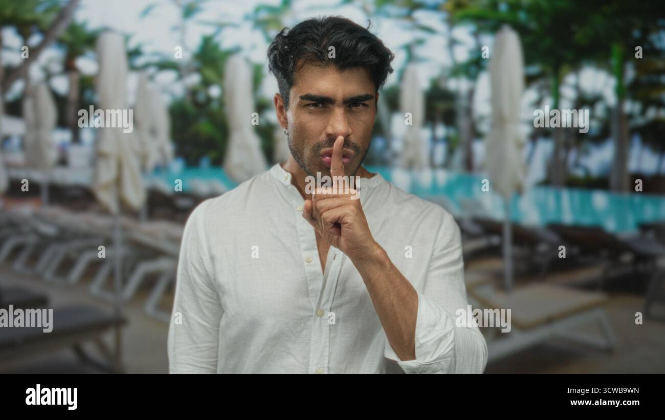 Giovane uomo in camicia di lino bianca in piedi a bordo piscina dell'edificio del resort con le dita alle labbra per il silenzio; segretezza. Foto Stock
