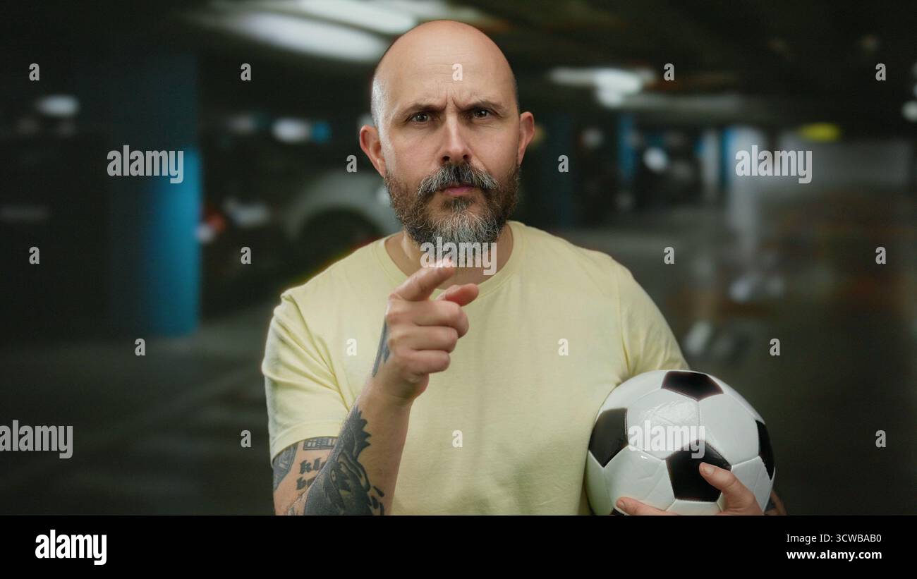 Uomo calvo con barba che tiene una palla da calcio in un parcheggio interno, che indica con serietà e mostra un braccio tatuato. Foto Stock