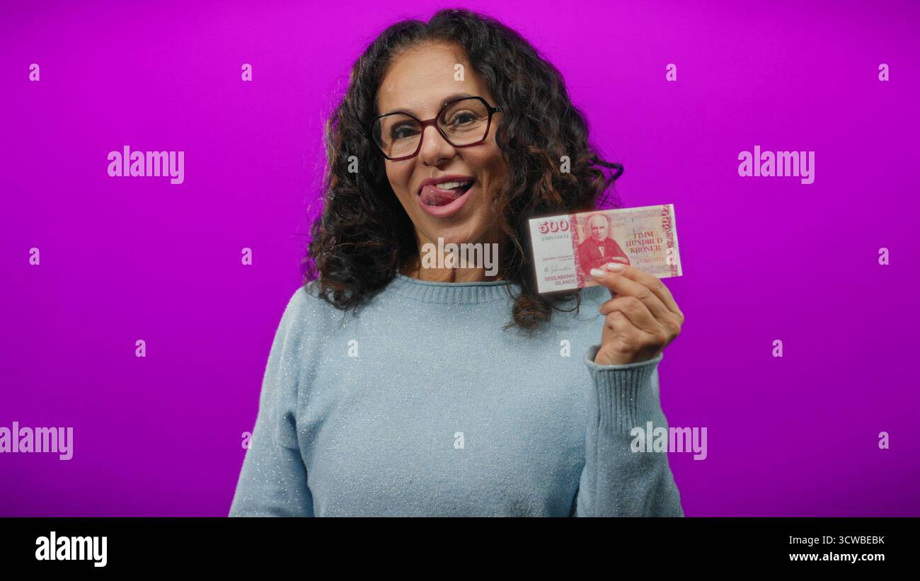 Donna di mezza età che tiene in mano la corona islandese sorridente su un vivace sfondo viola. Foto Stock