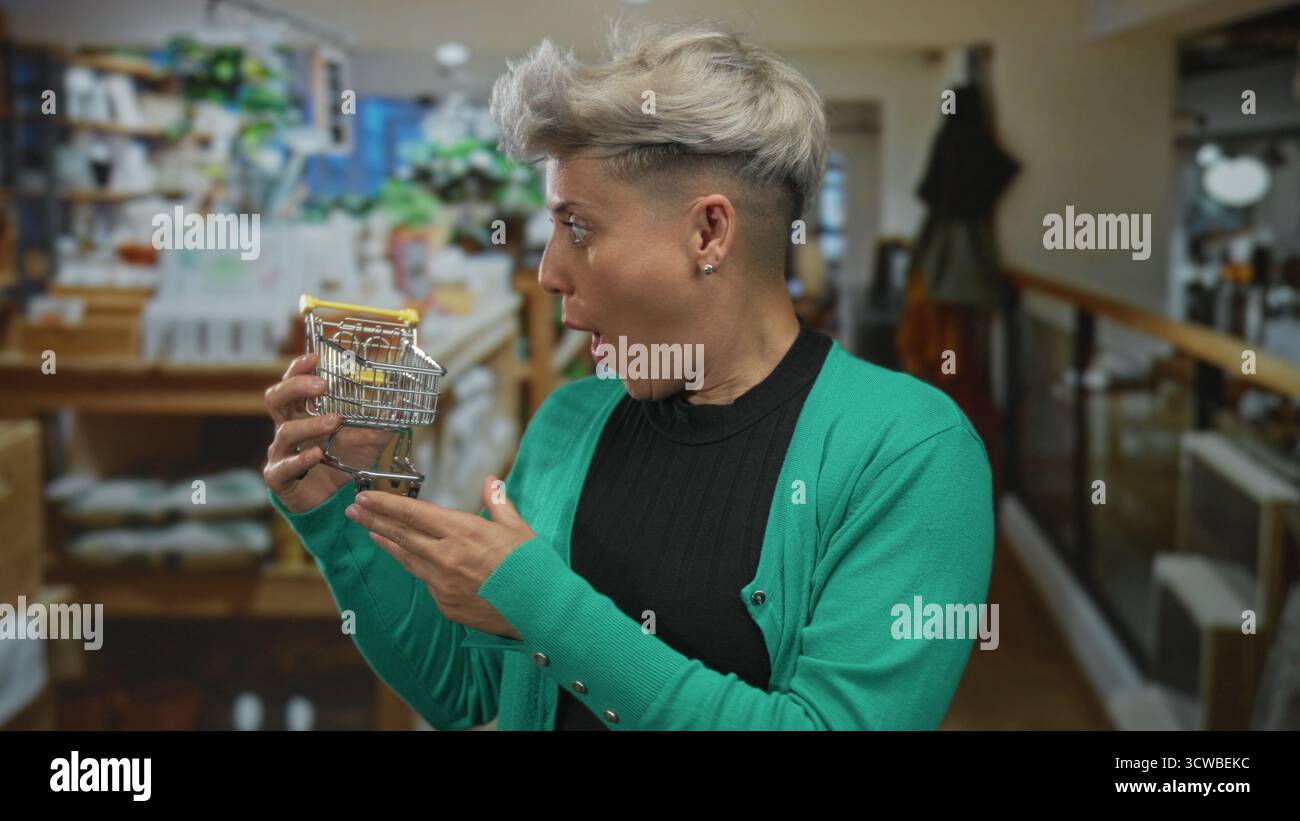 La donna dai capelli corti grigi tiene il carrello della spesa in miniatura con entrambe le mani a livello del petto nell'edificio; sorpresa. Foto Stock
