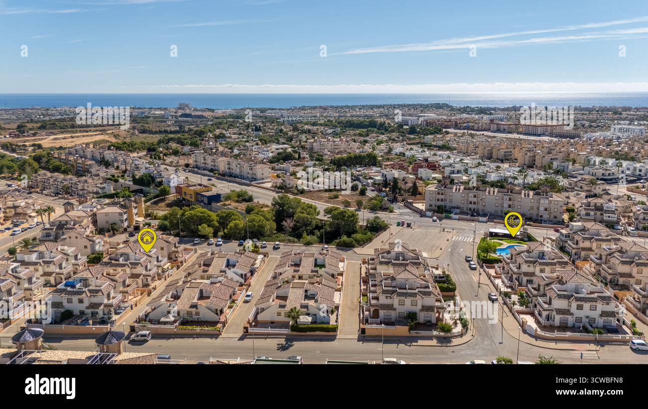 Vista aerea di un quartiere residenziale in stile mediterraneo vicino alla costa, con icone chiare che segnano una casa e una piscina sotto un cielo blu. Foto Stock