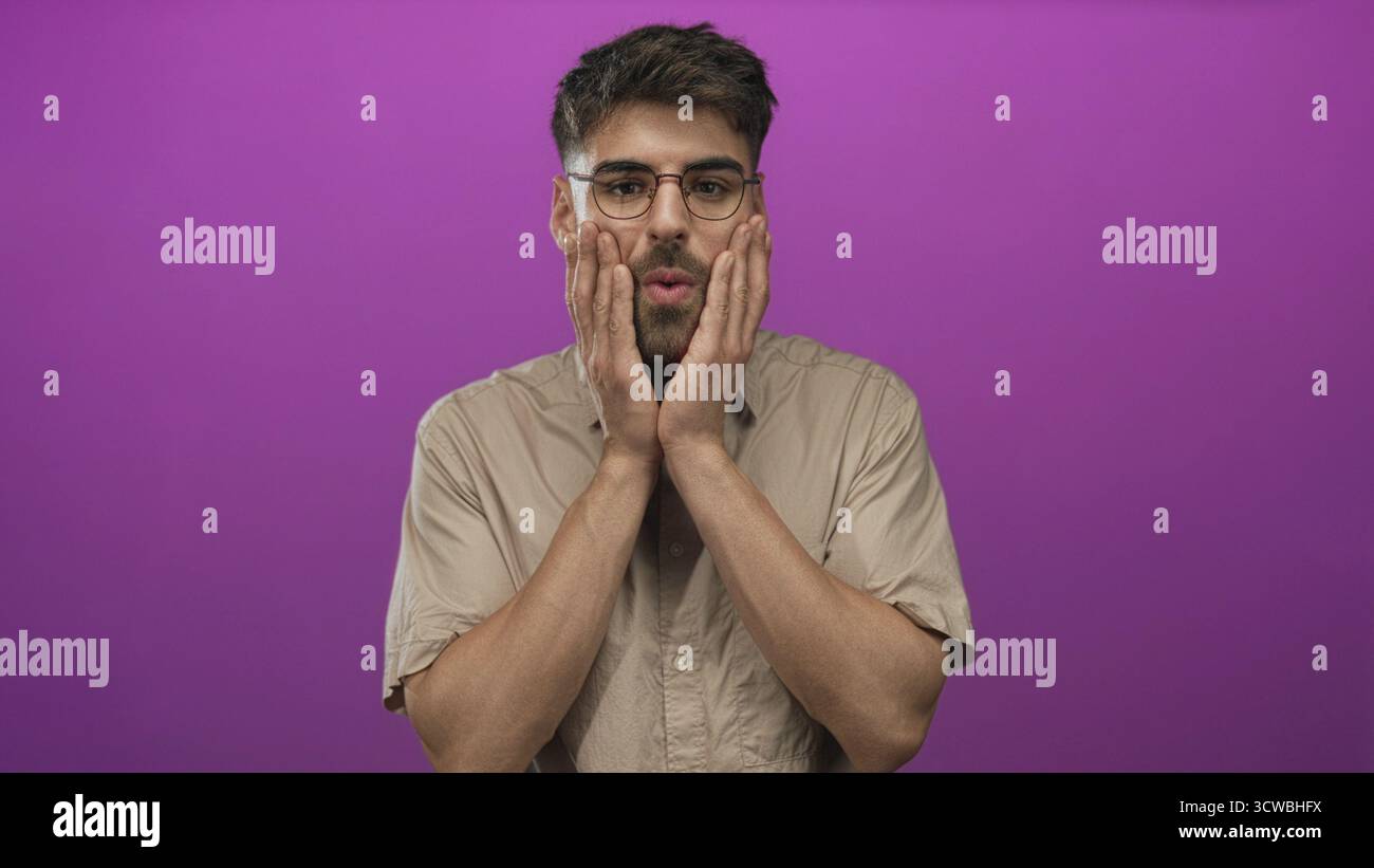 L'uomo che indossa occhiali e camicia beige preme entrambe le mani sulle guance con la bocca aperta e le sopracciglia in studio viola; sorpresa. Foto Stock
