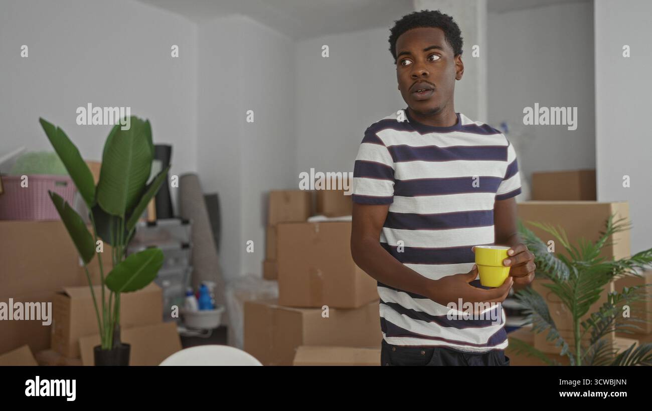 L'uomo tiene la tazza vicino a scatole non imballate e a una pianta in vaso mentre si appoggia al laptop nell'edificio; ha concentrato la pianificazione di nuovi inizi. Foto Stock