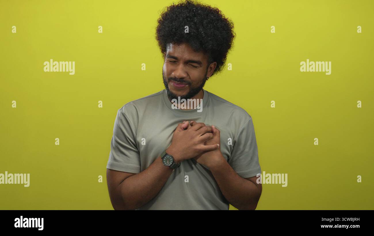 Uomo che tiene il petto davanti al muro giallo con un'espressione dolorosa, indossa una camicia grigia, capelli ricci e guarda, trasmettendo emozione e solitudine. Foto Stock