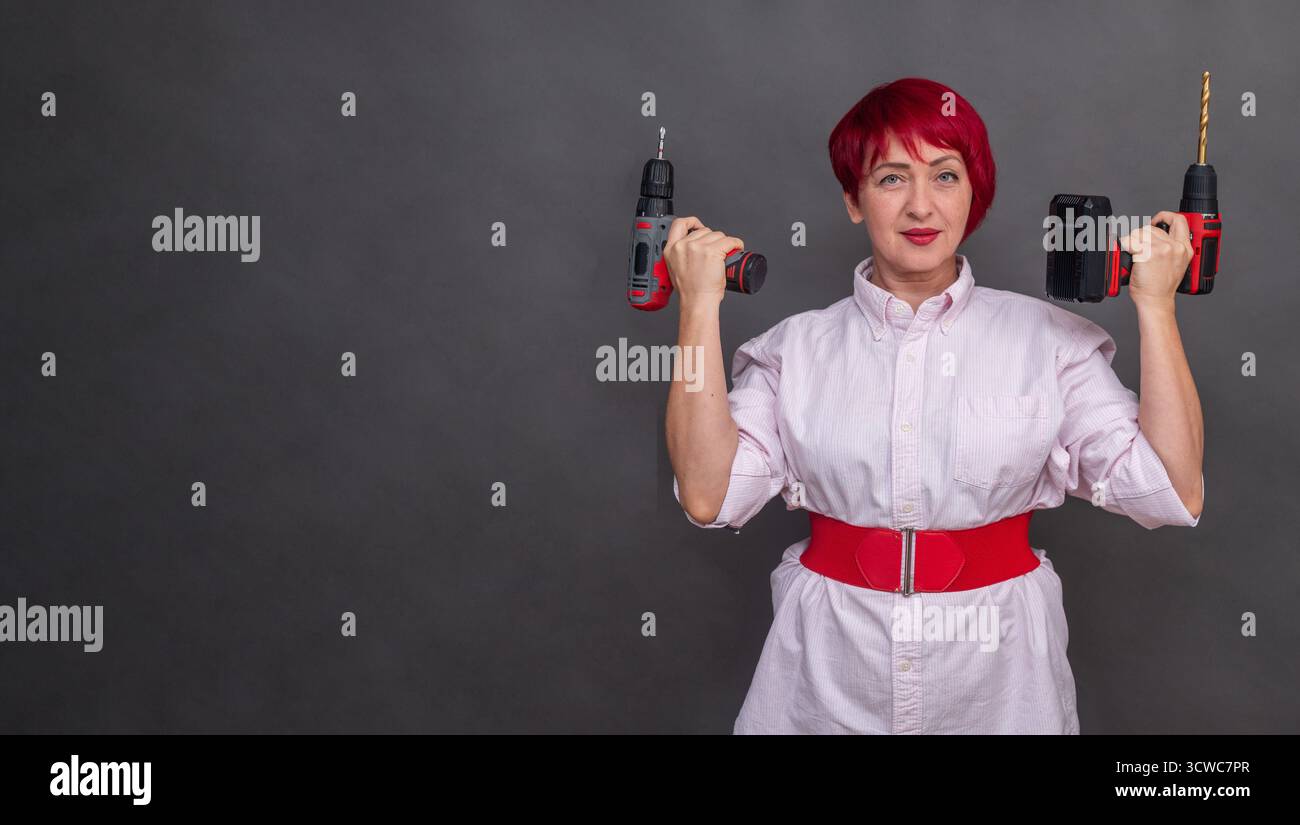 Donna sicura di sé con i capelli rossi può contenere due cacciaviti senza filo, pronti per lavori di costruzione. Gli strumenti professionali su sfondo grigio accentuano la Foto Stock