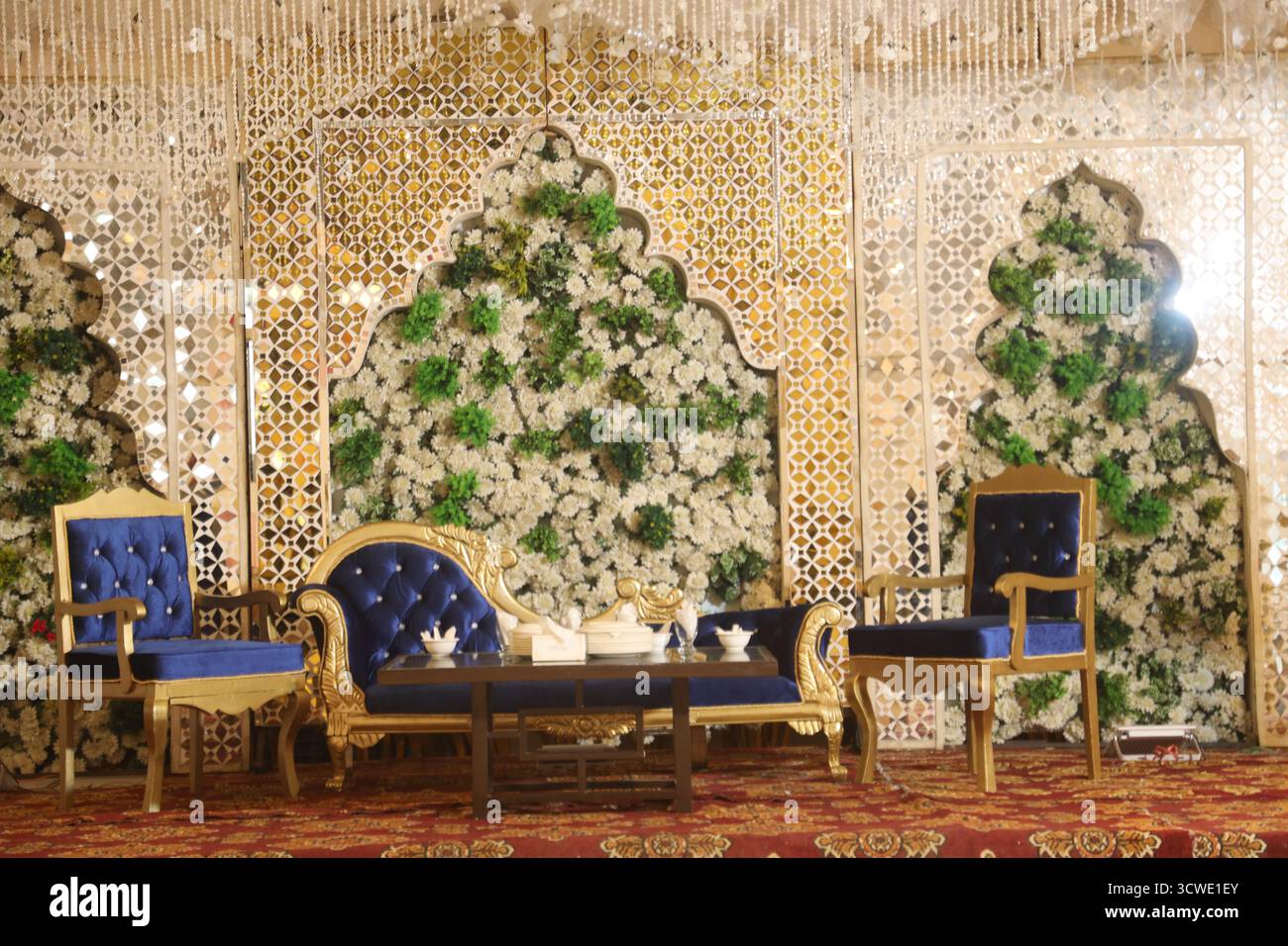 Arredi lussuosi per il palco di nozze con mobili Royal Blue e Gold, parete con sfondo floreale, eleganti pannelli a specchio e illuminazione a lampadario per ricevimenti Foto Stock