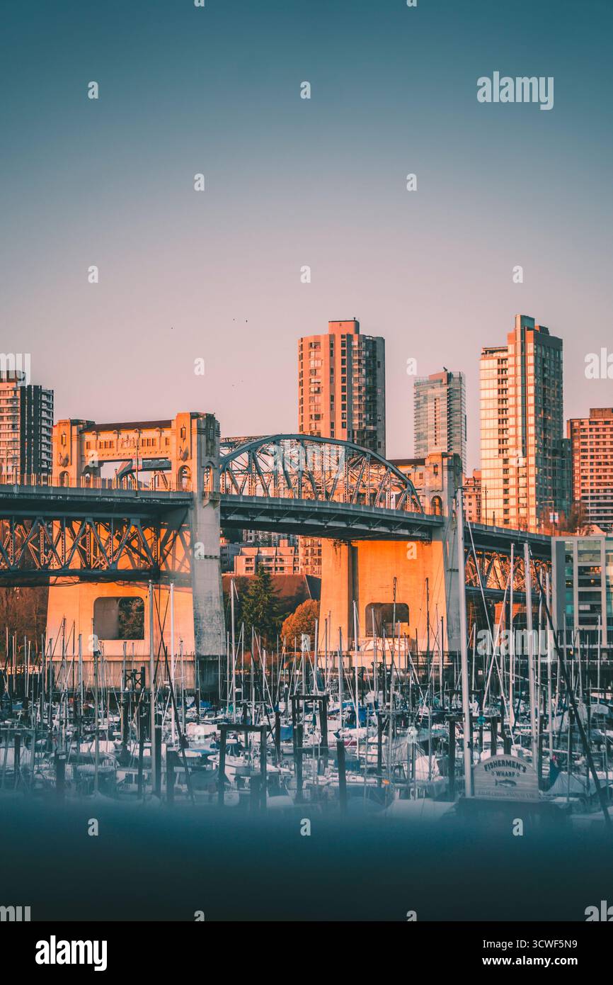 Un tramonto proietta una luce calda sulle barche nel porticciolo con uno sfondo di alti edifici cittadini e un ponte storico a Vancouver. Foto Stock