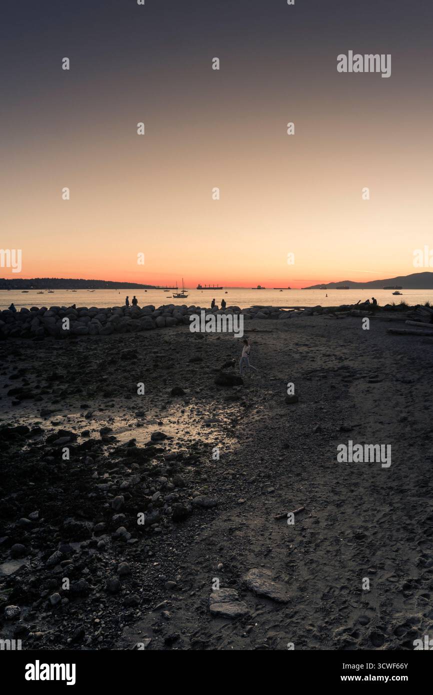 Le persone camminano lungo la riva mentre il sole tramonta sull'acqua, creando una colorata scena crepuscolare con sagome all'orizzonte. Foto Stock