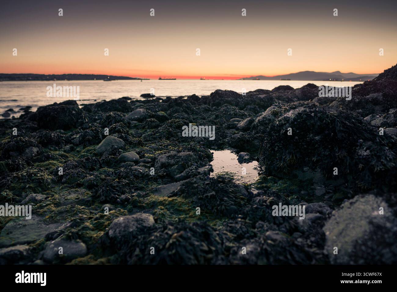 Le onde si infrangono dolcemente sulle coste rocciose mentre il sole tramonta, dipingendo il cielo con colori caldi sul mare calmo. Le barche sono visibili in lontananza. Foto Stock