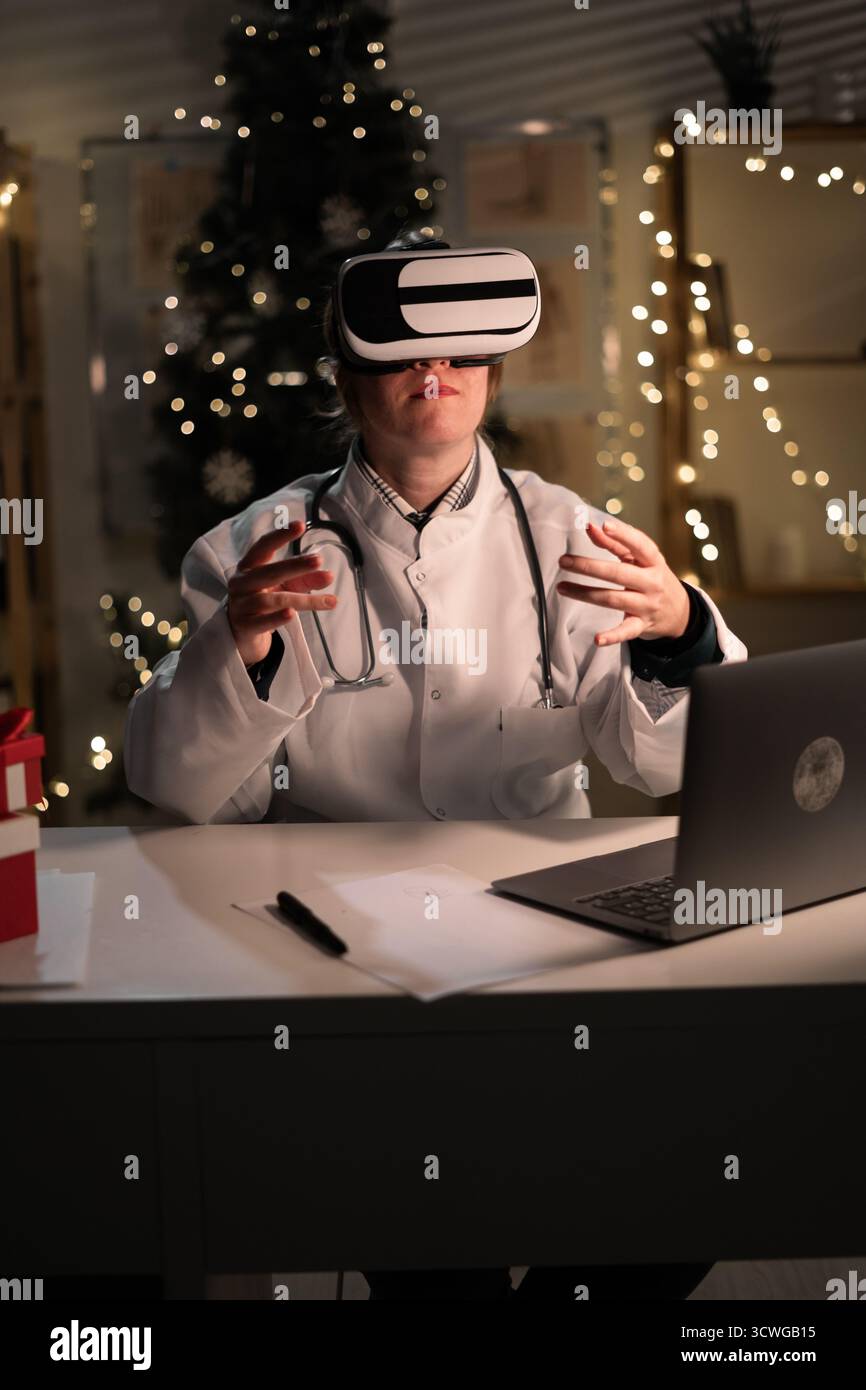 La giovane dottoressa indossa il cappello di Babbo Natale e gli occhiali vr che guardano la realtà metaversa durante il turno di lavoro la sera della vigilia di Natale. Medicina, santo Foto Stock