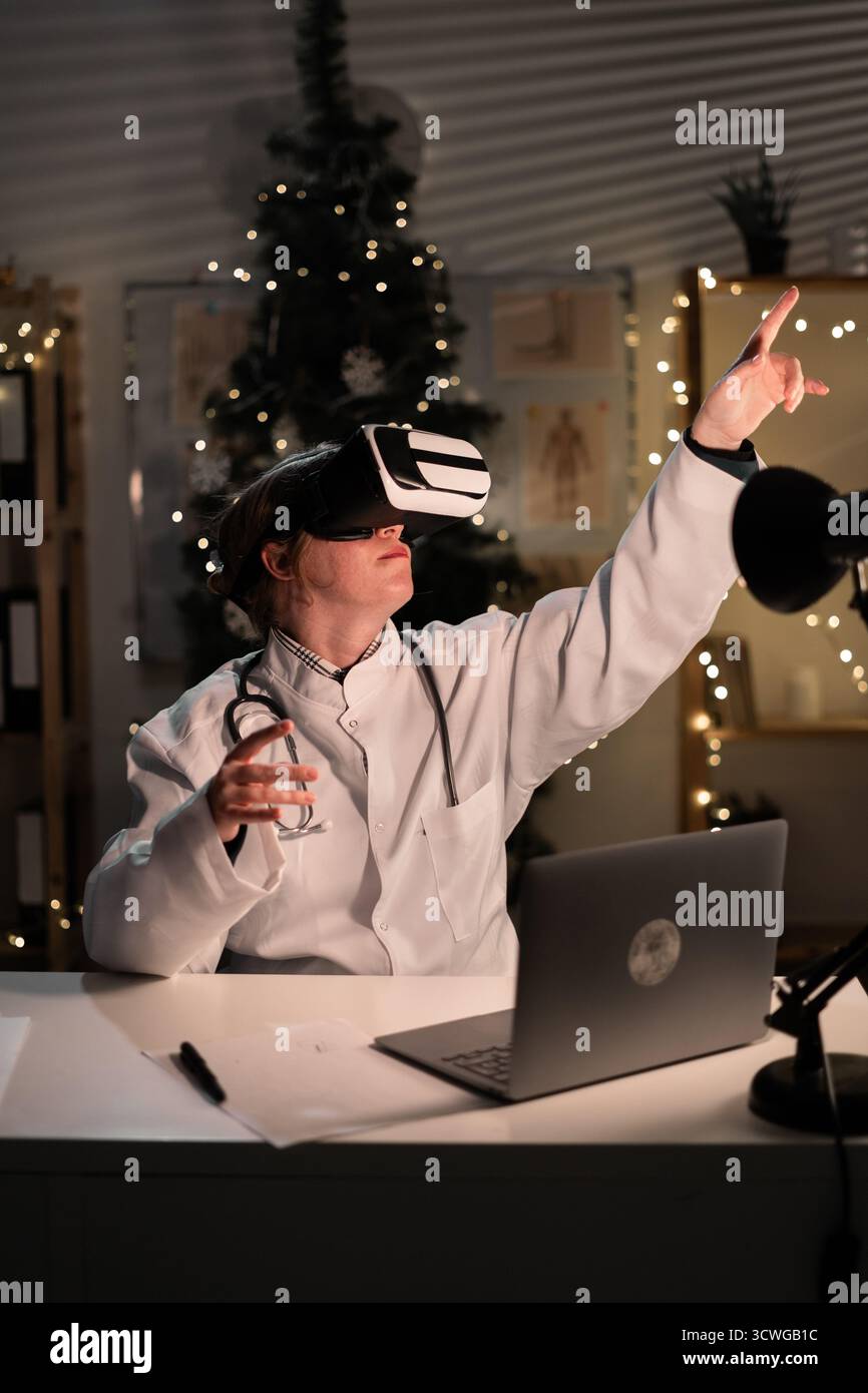 La giovane dottoressa indossa il cappello di Babbo Natale e gli occhiali vr che guardano la realtà metaversa durante il turno di lavoro la sera della vigilia di Natale. Medicina, vacanze, Foto Stock