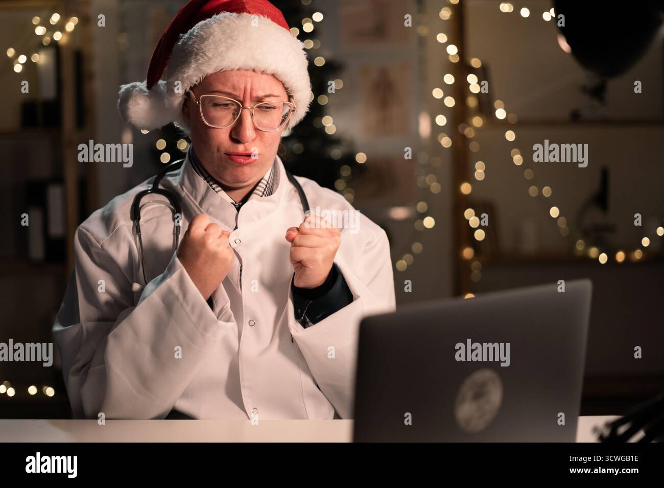 La giovane dottoressa indossa il cappello di Babbo Natale con un computer portatile che tiene i pugni durante il turno di notte la vigilia di Natale. Concetto di medicina, vacanze Foto Stock