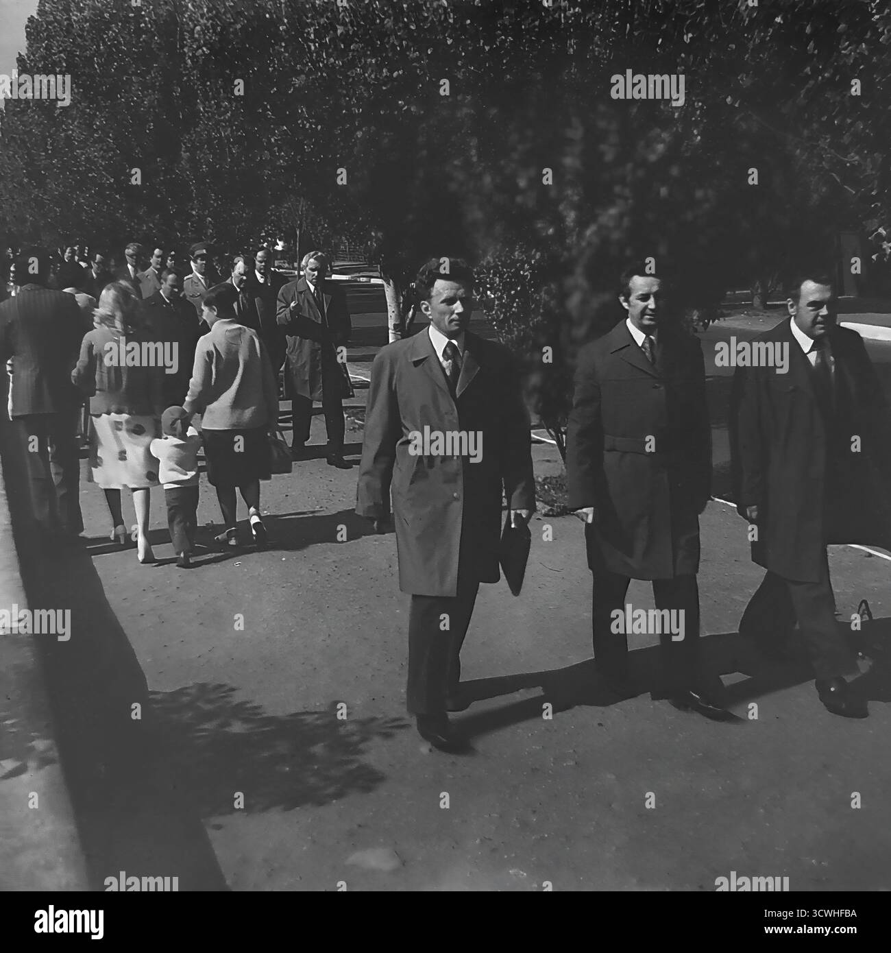 Un gruppo di uomini sovietici ben vestiti, probabilmente funzionari o delegati, cammina lungo un viale alberato soleggiato in via Karl Marx (ora via Tsentralna) a Sloviansk, RSS Ucraina. Questa candida foto in bianco e nero degli anni '1970 cattura l'atmosfera della vita pubblica durante l'era Brezhnev, con forti contrasti tra luce e ombra. La scena è una tipica rappresentazione di una processione o di un raduno ufficiale in una città provinciale dell'URSS Foto Stock