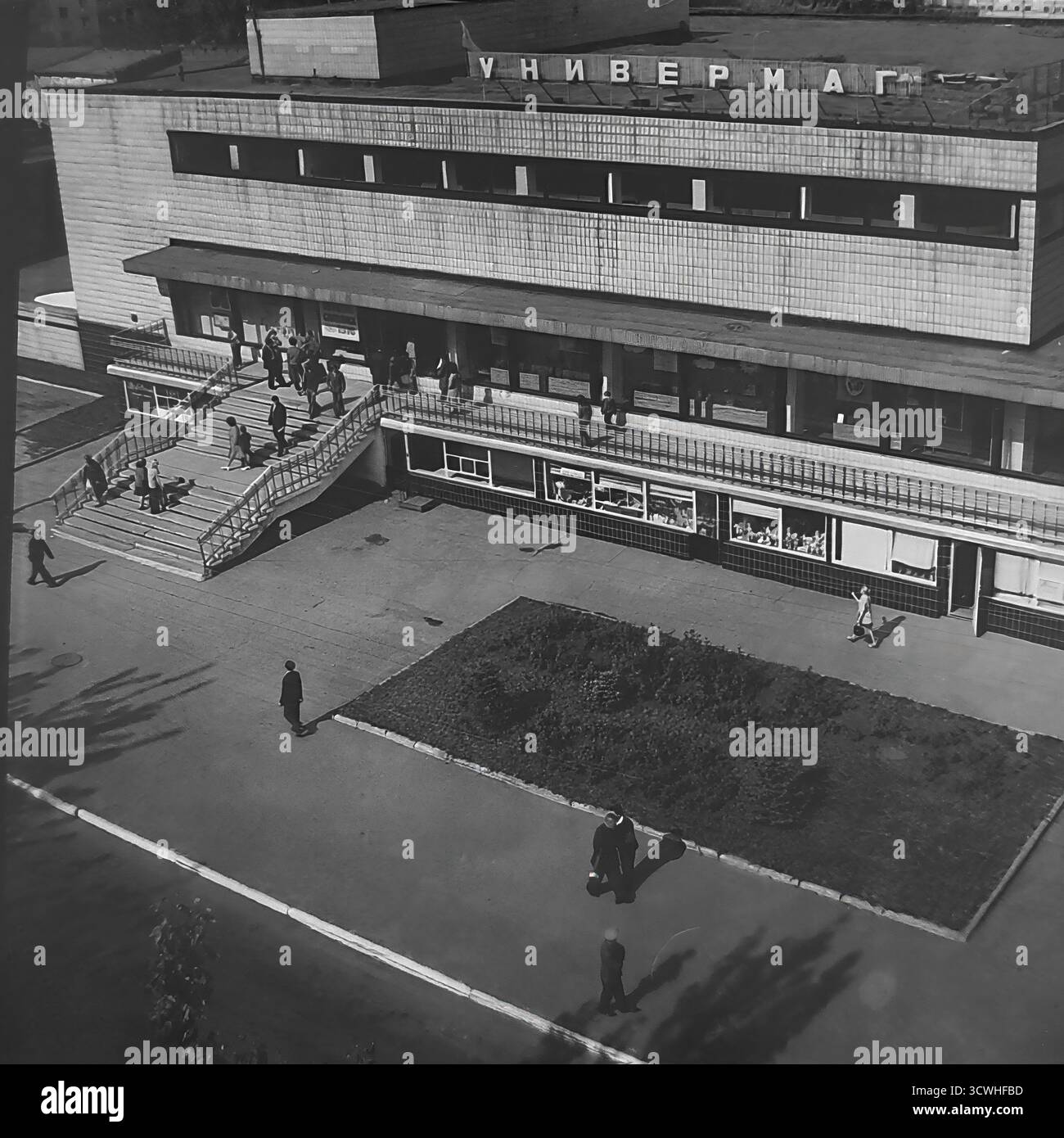 Una vista ad alto angolo cattura il grande magazzino centrale ("Univermag") e la vivace Piazza della Rivoluzione d'ottobre (ora Piazza Soborna) a Sloviansk, RSS Ucraina, durante gli anni '1970 L'edificio è un classico esempio di architettura modernista sovietica, che funge da principale centro commerciale della città. I pedoni passeggiano lungo gli ampi marciapiedi e la piazza, mostrando la vita quotidiana e l'ambiente urbano di una tipica città provinciale dell'URSS durante l'era Brezhnev. Questa foto in bianco e nero evoca un forte senso di nostalgia per il periodo Foto Stock