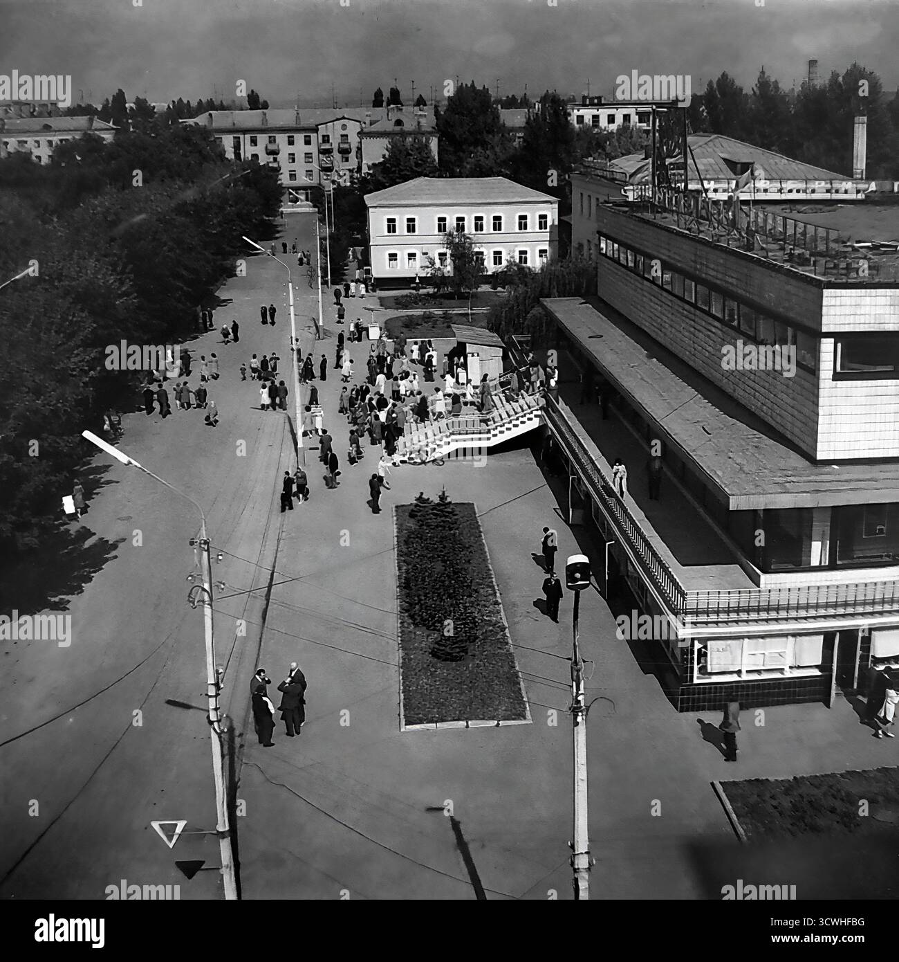 Una vista ad alto angolo cattura il grande magazzino centrale ("Univermag") e la vivace Piazza della Rivoluzione d'ottobre (ora Piazza Soborna) a Sloviansk, RSS Ucraina, durante gli anni '1970 L'edificio è un classico esempio di architettura modernista sovietica, che funge da principale centro commerciale della città. I pedoni passeggiano lungo gli ampi marciapiedi e la piazza, mostrando la vita quotidiana e l'ambiente urbano di una tipica città provinciale dell'URSS durante l'era Brezhnev. Questa foto in bianco e nero evoca un forte senso di nostalgia per il periodo Foto Stock