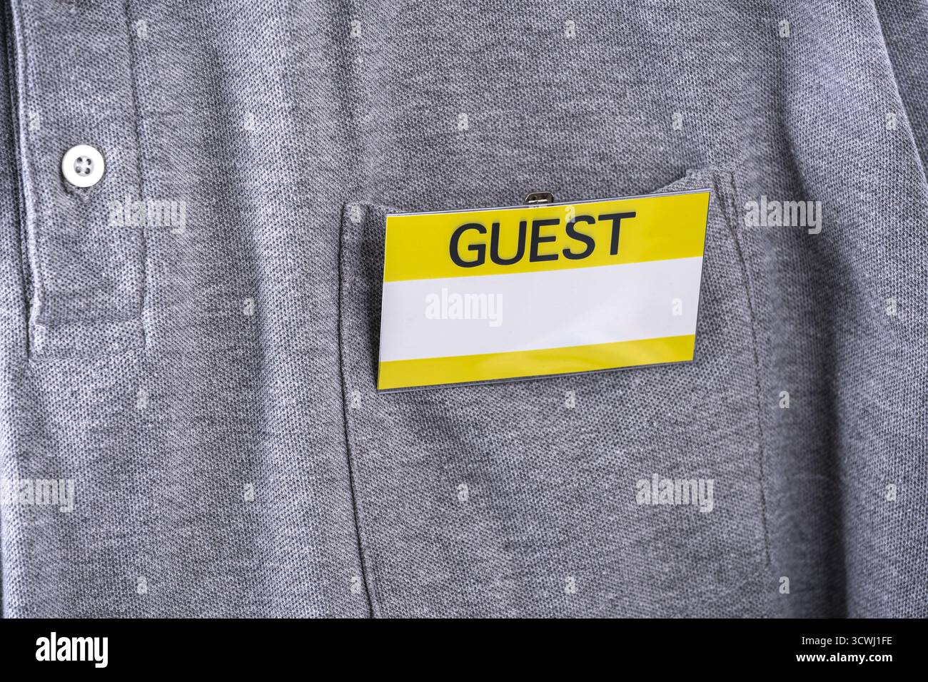 Un distintivo per gli ospiti con bordo giallo è fissato alla tasca della camicia grigia, a indicare lo stato dei visitatori. il distintivo si distingue dal tessuto, suggerendo un aspetto formale Foto Stock
