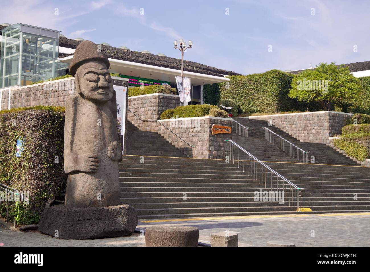 Un tradizionale guardiano di pietra hareubang dol si erge sui gradini del Museo del Folklore e di storia naturale di Jeju, con striscioni e pareti rivestite di edera Foto Stock