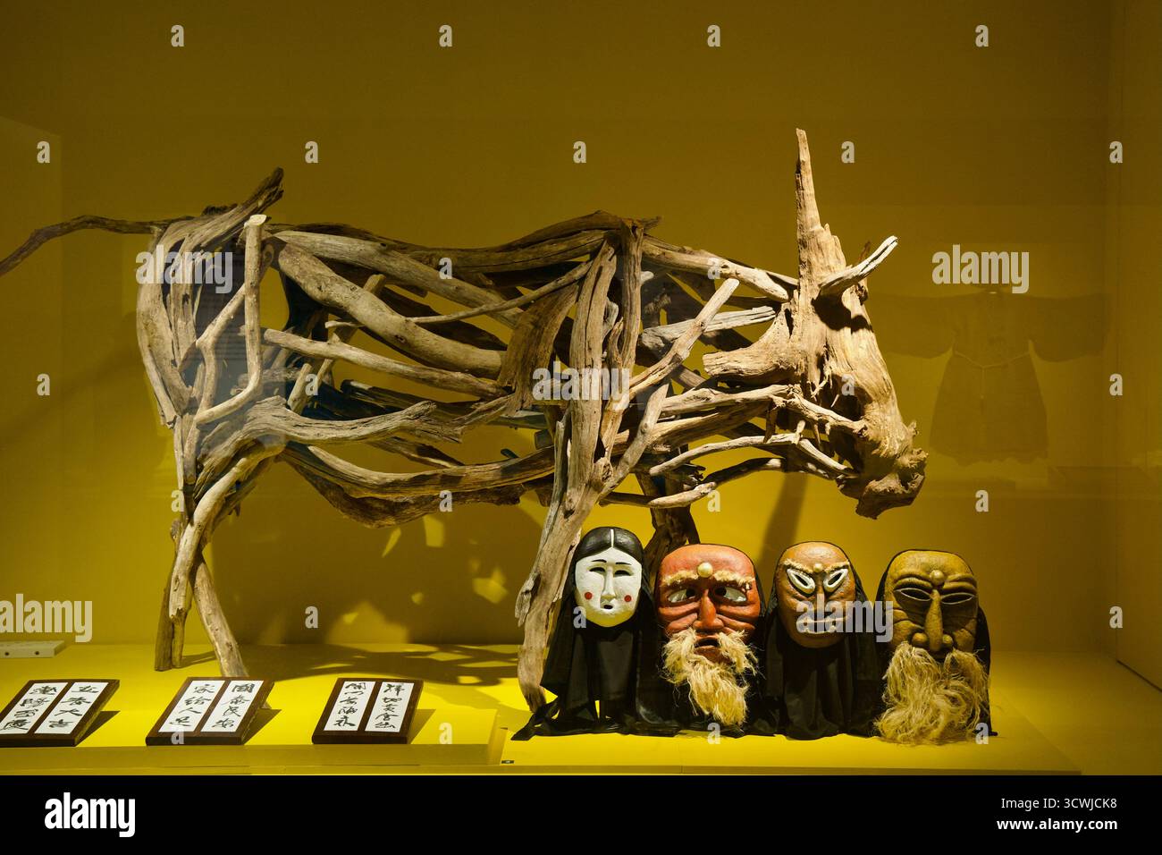 Una forma di bue scheletrico costruita da legno di mare si erge dietro quattro maschere scolpite per festival in una vignetta della galleria del Museo del Folklore e di storia naturale di Jeju, Foto Stock