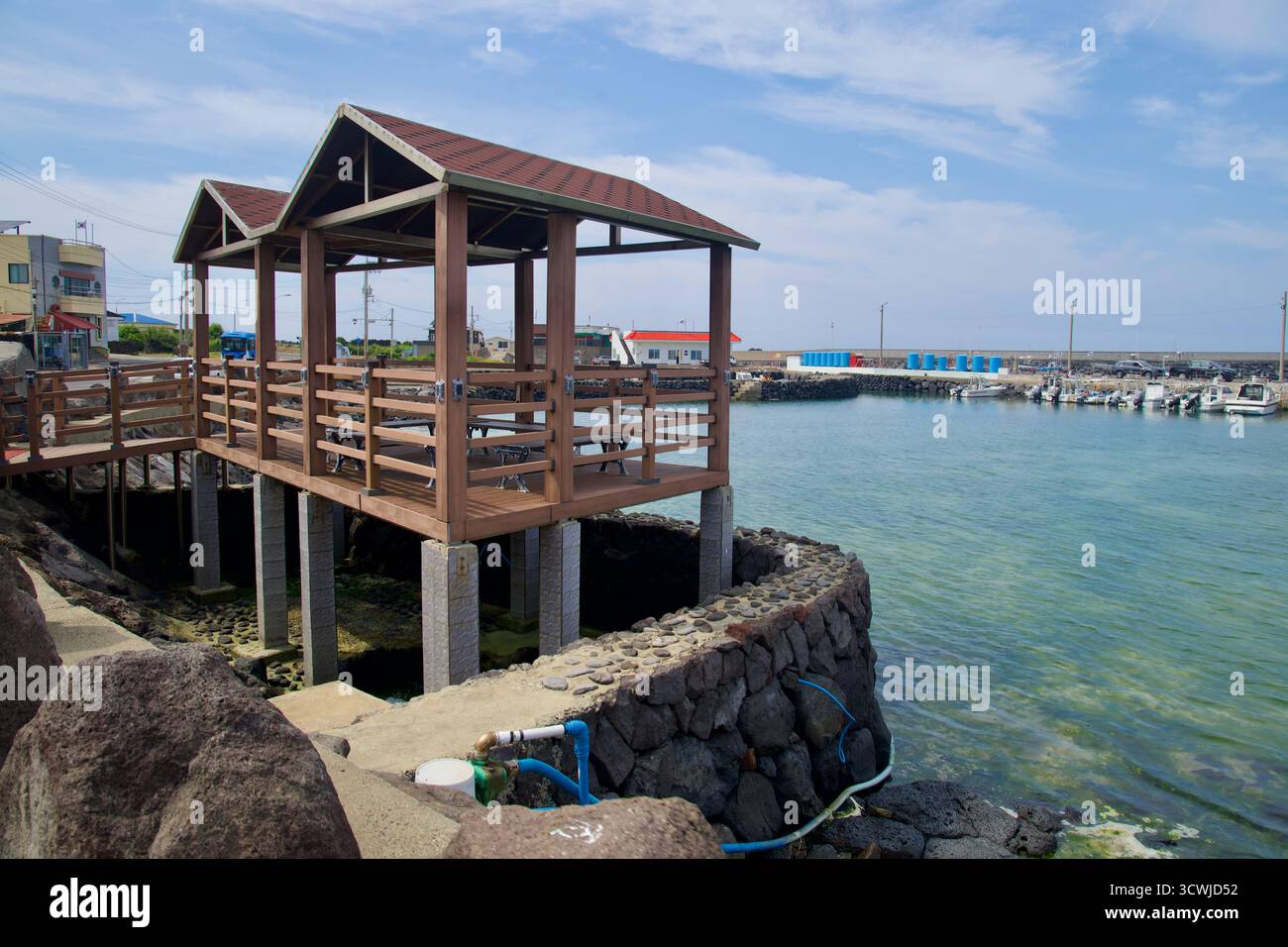Un padiglione in legno sul lungomare su palafitte si affaccia sul porto di Beollang, con vasche di stoccaggio blu e barche ormeggiate che costeggiano il porto sotto nuvole sparse. Foto Stock
