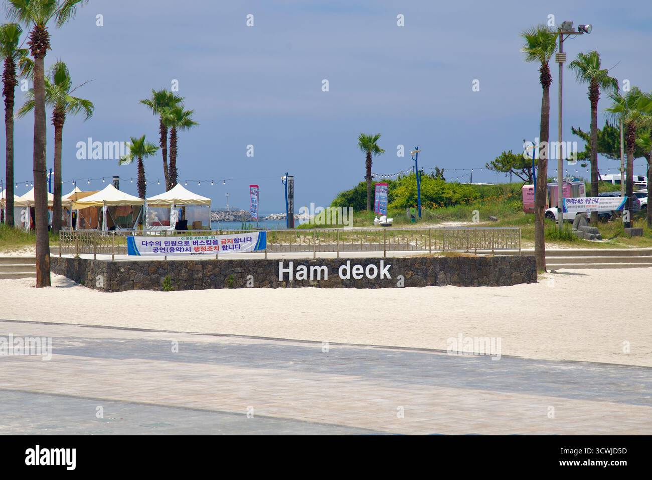 Una bassa parete di basalto con un cartello con il nome Hamdeok si affaccia sul lungomare con palme, tende da festival e luci costiere sulla sabbia di Hamdeok Seou Foto Stock