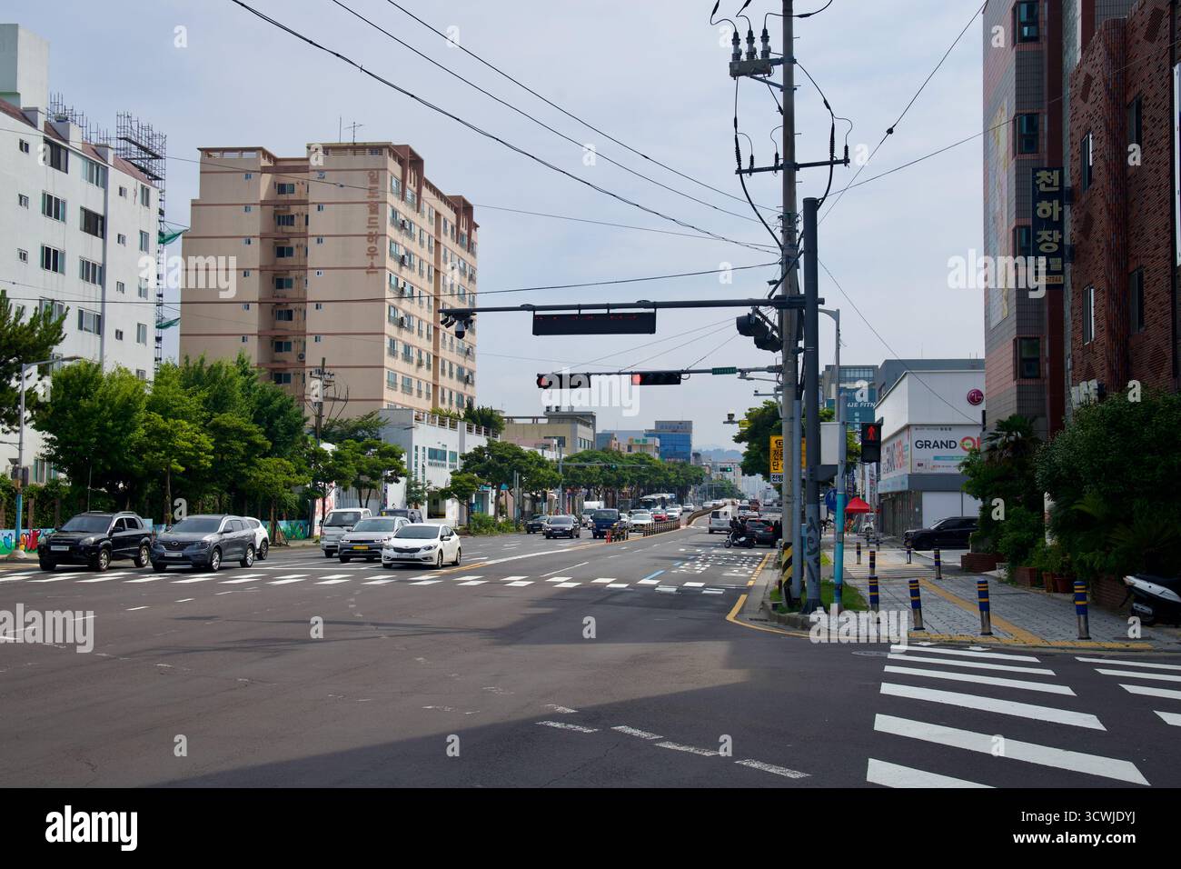 Il traffico attende a un semaforo rosso lungo una strada principale nel quartiere di Hwabuk, fiancheggiata da appartamenti di media altezza, negozi e linee di servizio che conducono a Jeju's. Foto Stock