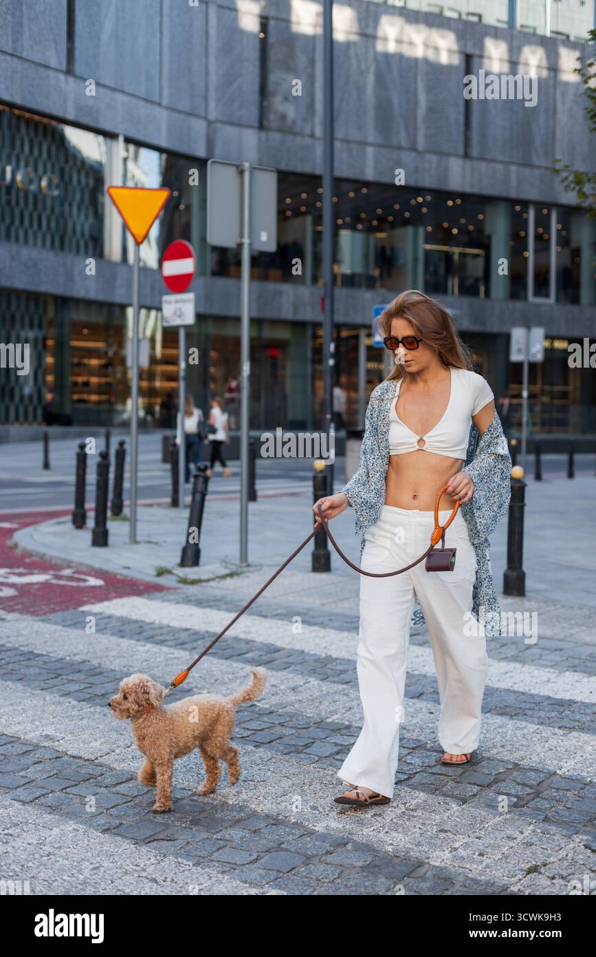 Donna alla moda che cammina il suo cane su Una strada della città. Vestita con pantaloni bianchi e Un Crop Top, trasuda fiducia e stile urbano moderno. Momento accogliente W Foto Stock