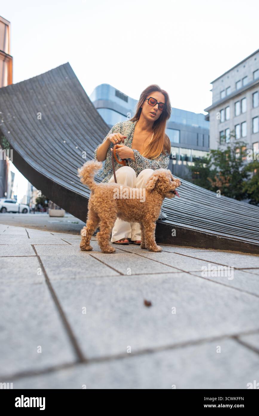Un momento di stile urbano con Una donna e il suo barboncino che si godono Una pausa tranquilla. Accarezza il barboncino al guinzaglio, facendo una passeggiata e girando in Una piazza della città. Foto Stock