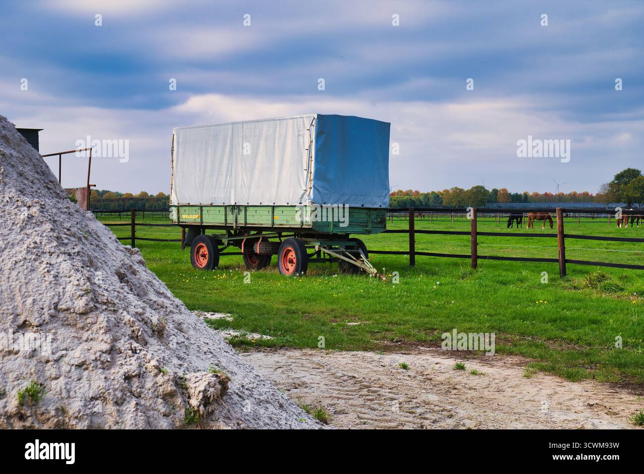 Rimorchio vintage Green Farm parcheggiato accanto al White Lime Pile in Grassy Field con recinzione in legno e cavalli da pascolo in lontananza sotto Blue Autumn Sky Foto Stock