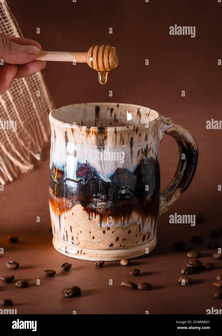 Un cucchiaio di miele che gocciola il miele in un caffè in ceramica, i chicchi di caffè versati in primo piano e fondo marrone Foto Stock