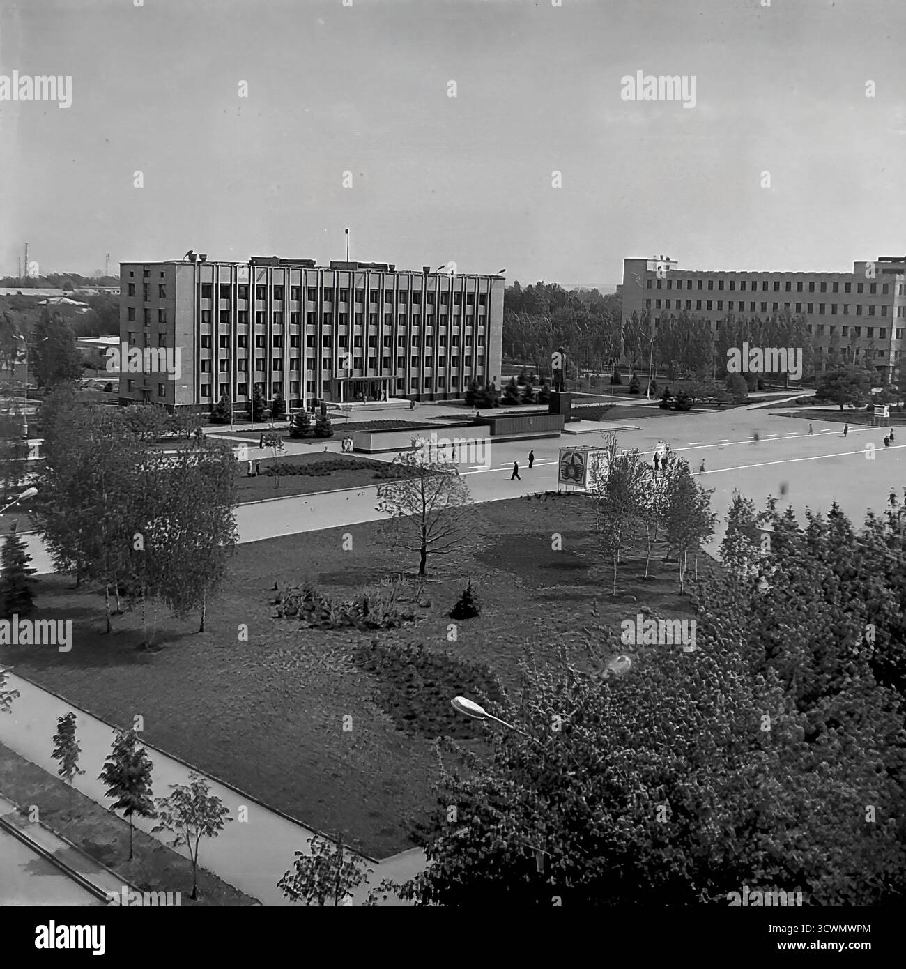 Una vista panoramica ad alto angolo cattura il principale cuore amministrativo di Sloviansk, la RSS Ucraina, negli anni '1980 La scena mostra la vasta Piazza della Rivoluzione d'ottobre (ora Piazza Soborna), dominata dall'edificio del Consiglio comunale in stile funzionalista e dal monumento centrale a Vladimir Lenin. I pedoni passeggiano attraverso lo spazio pubblico ben tenuto con la sua vegetazione paesaggistica e gli ampi sentieri. Questa foto in bianco e nero è un documento perfetto di pianificazione urbana sovietica, che mostra il centro ufficiale e ideologico di una tipica città provinciale durante la tarda URSS Foto Stock