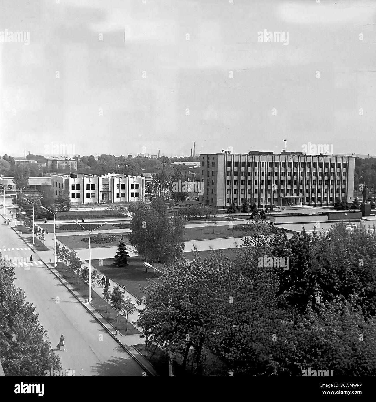 Una vista panoramica ad alto angolo cattura il principale cuore amministrativo di Sloviansk, la RSS Ucraina, negli anni '1980 La scena mostra la vasta Piazza della Rivoluzione d'ottobre (ora Piazza Soborna), dominata dall'edificio del Consiglio comunale in stile funzionalista e dal monumento centrale a Vladimir Lenin. I pedoni passeggiano attraverso lo spazio pubblico ben tenuto con la sua vegetazione paesaggistica e gli ampi sentieri. Questa foto in bianco e nero è un documento perfetto di pianificazione urbana sovietica, che mostra il centro ufficiale e ideologico di una tipica città provinciale durante la tarda URSS Foto Stock