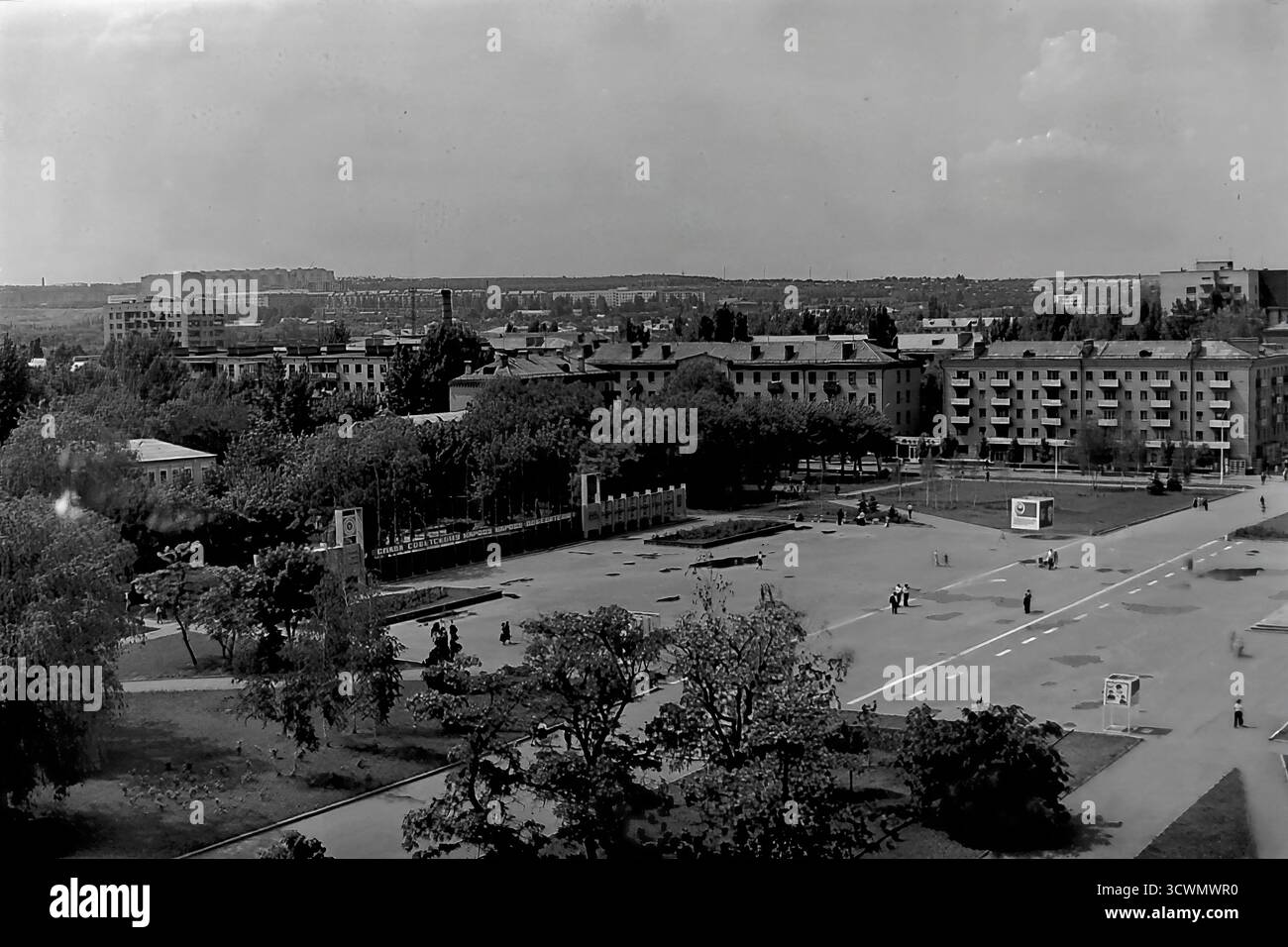 Una vasta vista panoramica cattura l'area verde del parco di Piazza della Rivoluzione d'ottobre (ora Piazza Soborna) a Sloviansk, RSS Ucraina, durante gli anni '1980 La scena mostra la classica pianificazione urbana sovietica, con un Public Honor Board in stile modernista ('Doska poeta'), ampi sentieri asfaltati e spazi verdi paesaggistici. Alcuni pedoni passeggiano attraverso l'immensa e tranquilla piazza. Sullo sfondo, un vasto paesaggio urbano con condomini e distanti fabbriche di fumo completa l'immagine di una tipica città provinciale della tarda URSS Foto Stock