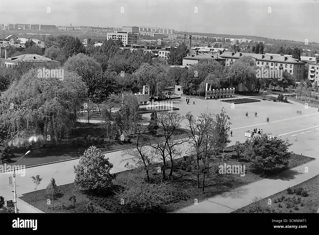 Una vasta vista panoramica cattura l'area verde del parco di Piazza della Rivoluzione d'ottobre (ora Piazza Soborna) a Sloviansk, RSS Ucraina, durante gli anni '1980 La scena mostra la classica pianificazione urbana sovietica, con un Public Honor Board in stile modernista ('Doska poeta'), ampi sentieri asfaltati e spazi verdi paesaggistici. Alcuni pedoni passeggiano attraverso l'immensa e tranquilla piazza. Sullo sfondo, un vasto paesaggio urbano con condomini e distanti fabbriche di fumo completa l'immagine di una tipica città provinciale della tarda URSS Foto Stock