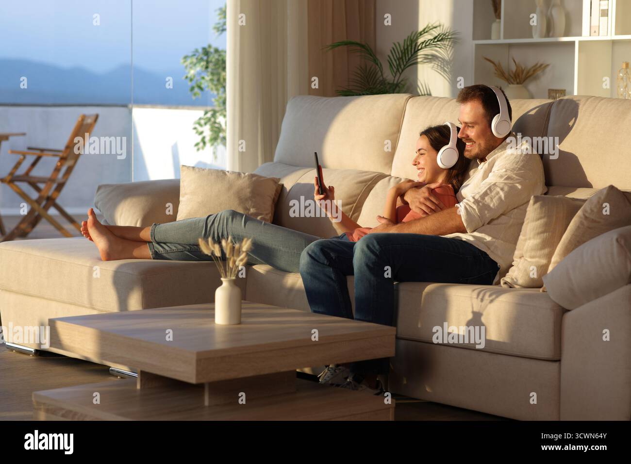Coppia felice che indossa le cuffie per condividere la musica dallo smartphone seduto sul divano di casa al tramonto Foto Stock