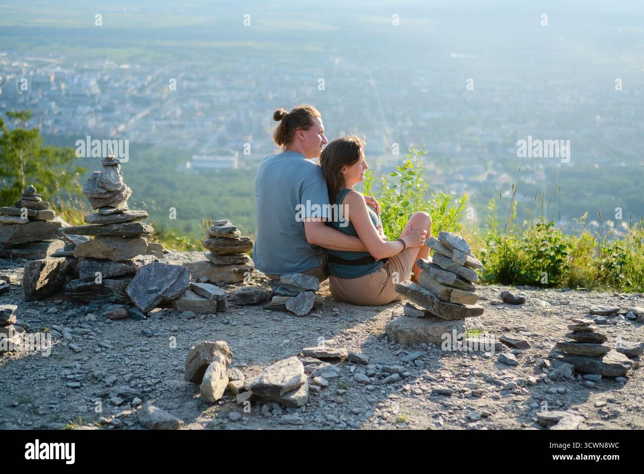 La giovane coppia seduto sul retro ammira il panorama mozzafiato della città dalla cima della montagna, con cairns in pietra in primo piano. Perfetto per viaggi e viaggi d'amore. Foto Stock