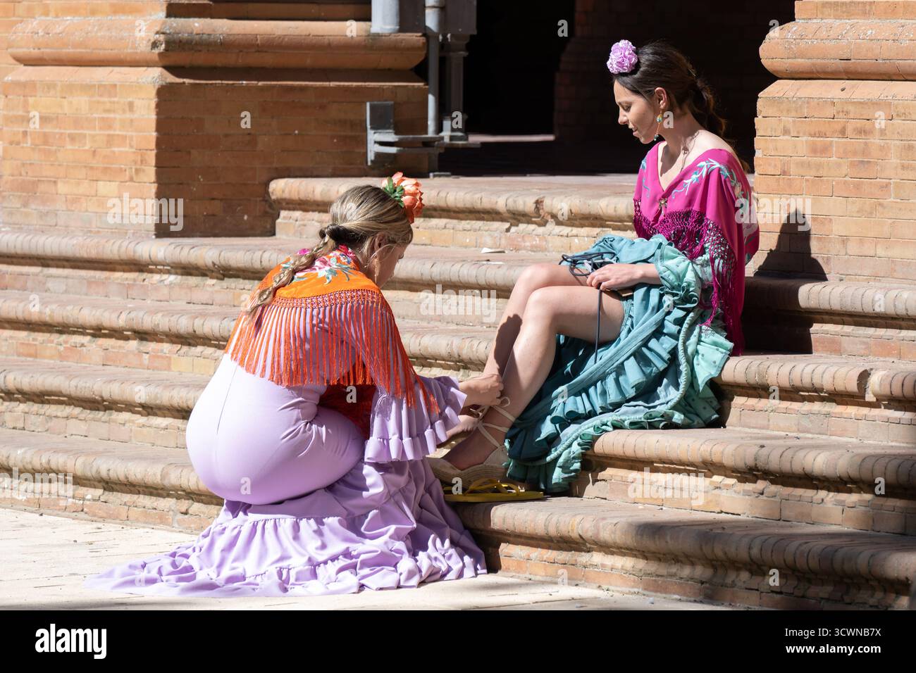 Siviglia, Spagna - 23 apr 23: Due donne in abiti tradizionali sedute su gradini, una che si adatta alle calzature dell'altra, colori vivaci, abbigliamento culturale Foto Stock