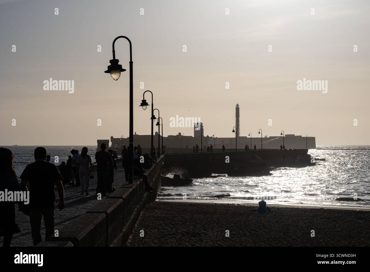 Figure sagomate che camminano lungo il molo di Cadice, Spagna al tramonto, con un faro sullo sfondo e onde morbide che riflettono la luce Foto Stock