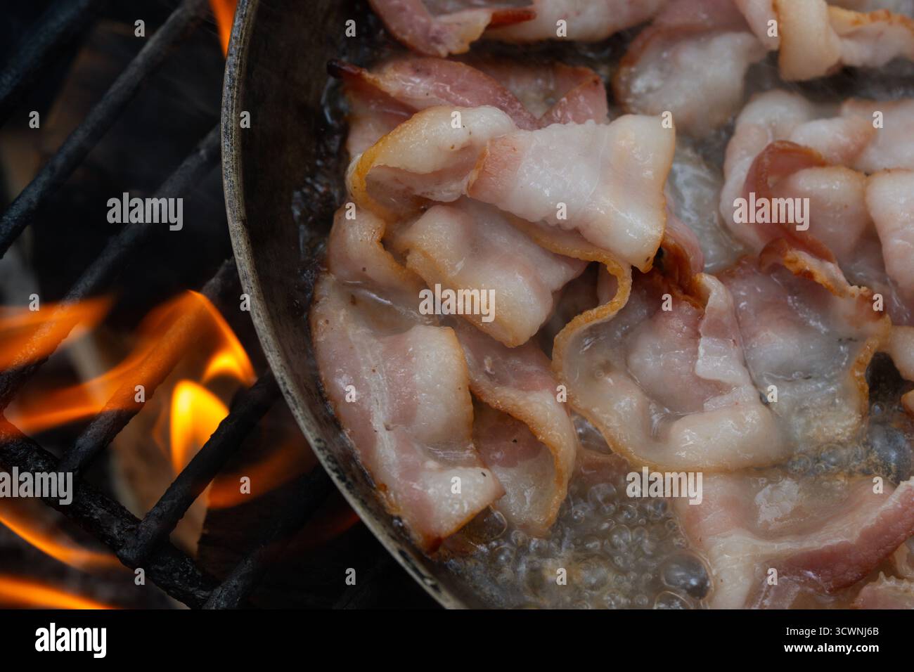 Bacon frizzante in una padella su un fuoco aperto che cucina all'aperto Foto Stock
