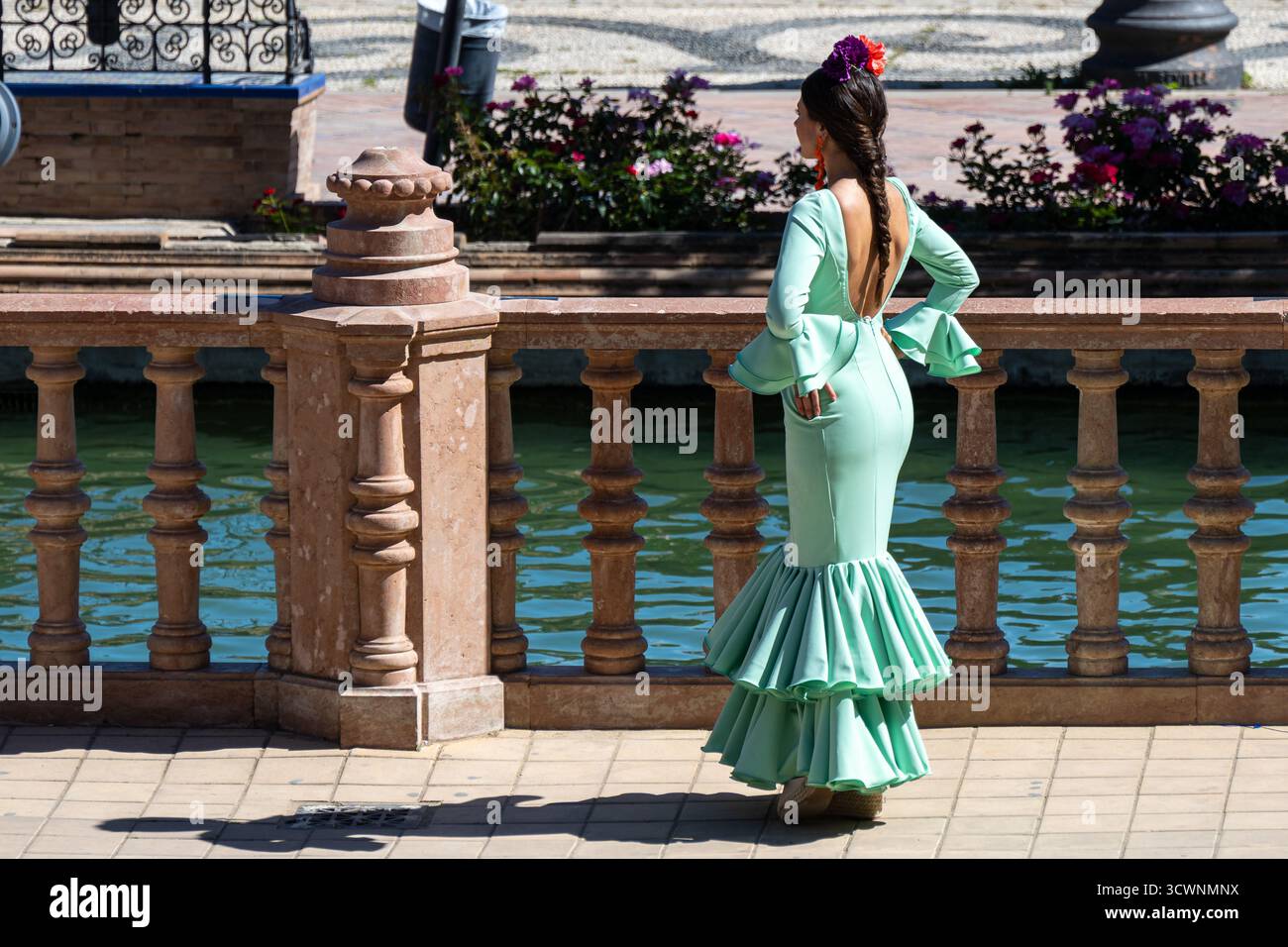 Siviglia, Spagna - 23 apr 23: Una donna con un abito verde menta con volant si erge su una ringhiera decorativa vicino ad un corpo d'acqua, circondata da fiori Foto Stock