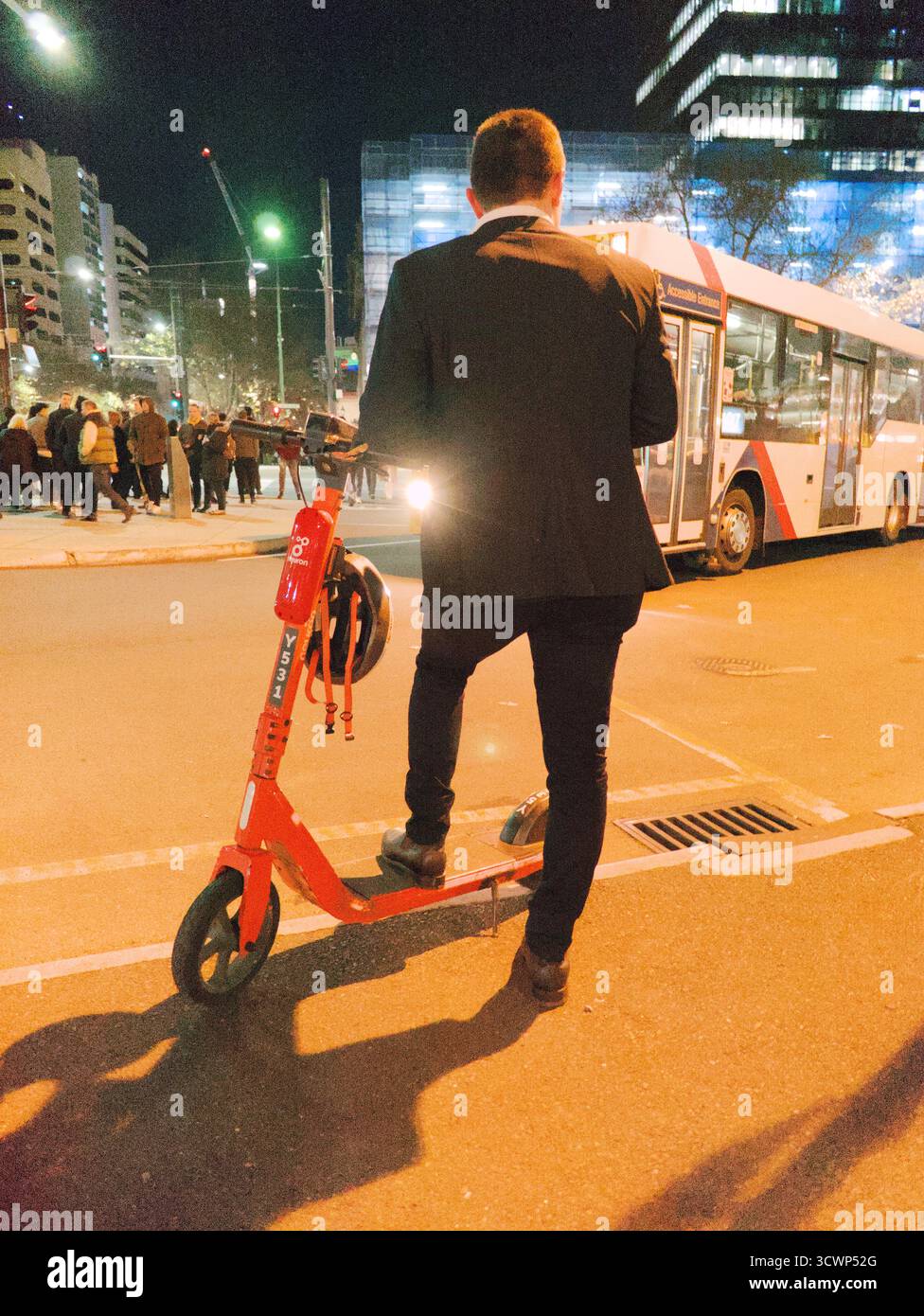 Moderno ufficio. Vista posteriore di un uomo d'affari su un voi e-scooter sotto lampioni dorati. Evoca sensazioni di velocità, lavoro tardi ed efficienza. Foto Stock